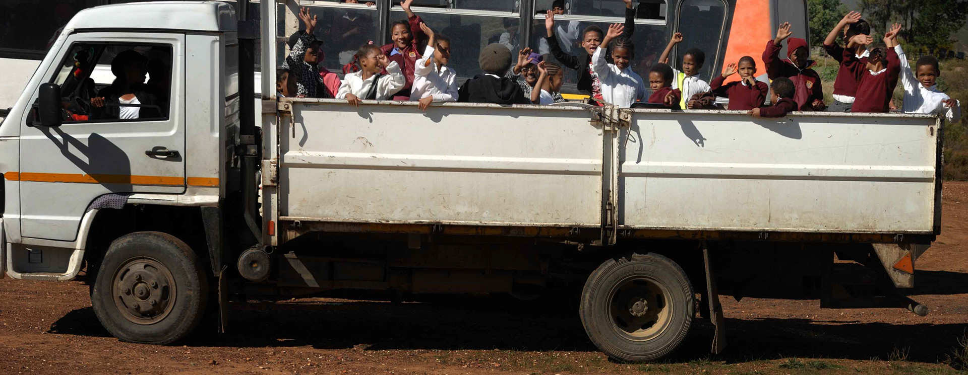 A white truck, filled with cheerful schoolchildren in uniforms, waves energetically from the back under a sunny sky. The mood is lively and joyful.
