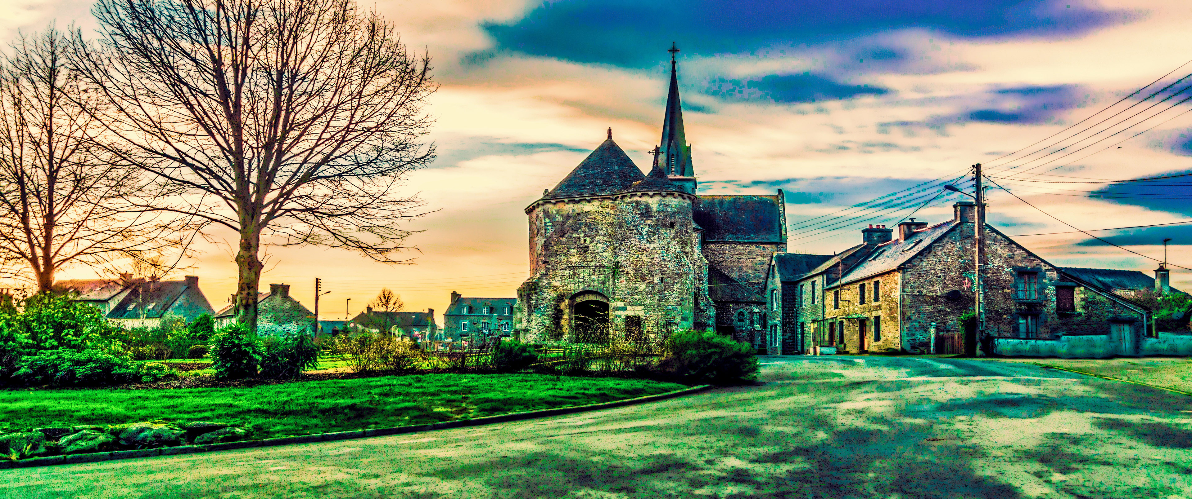 Medieval stone building with a conical roof and spire under a vibrant sunset sky. Leafless tree and green grass surround the tranquil village scene.