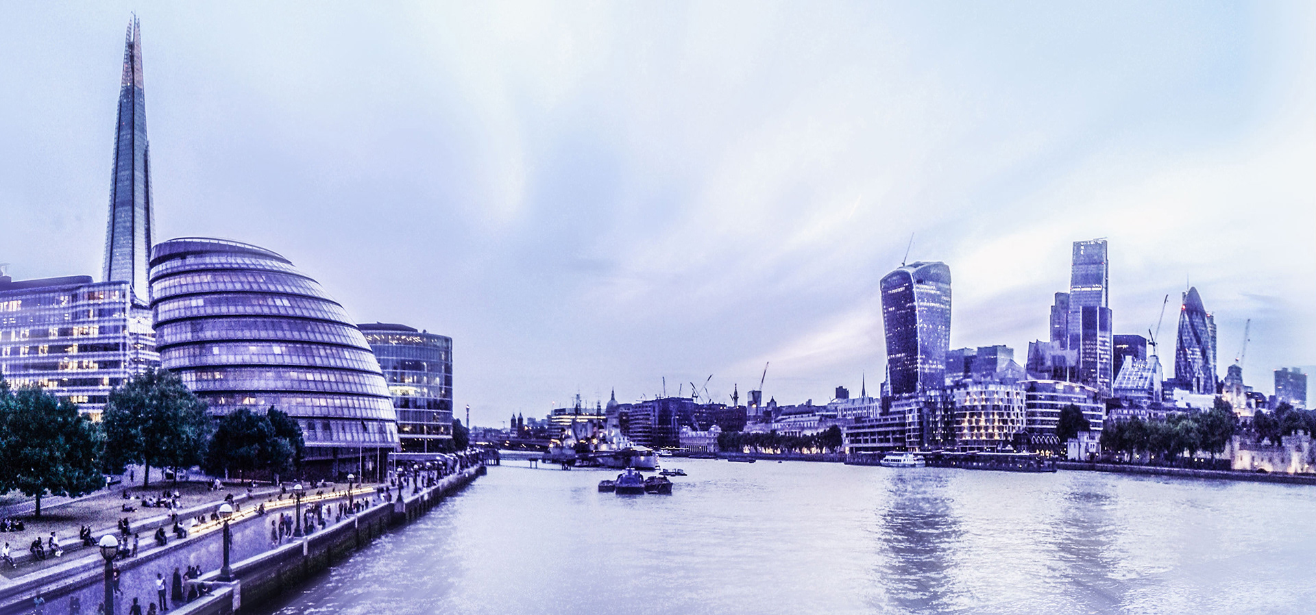 Panoramic view of London skyline at dusk, featuring illuminated modern buildings like The Shard and City Hall. The River Thames flows calmly, enhancing the serene atmosphere.