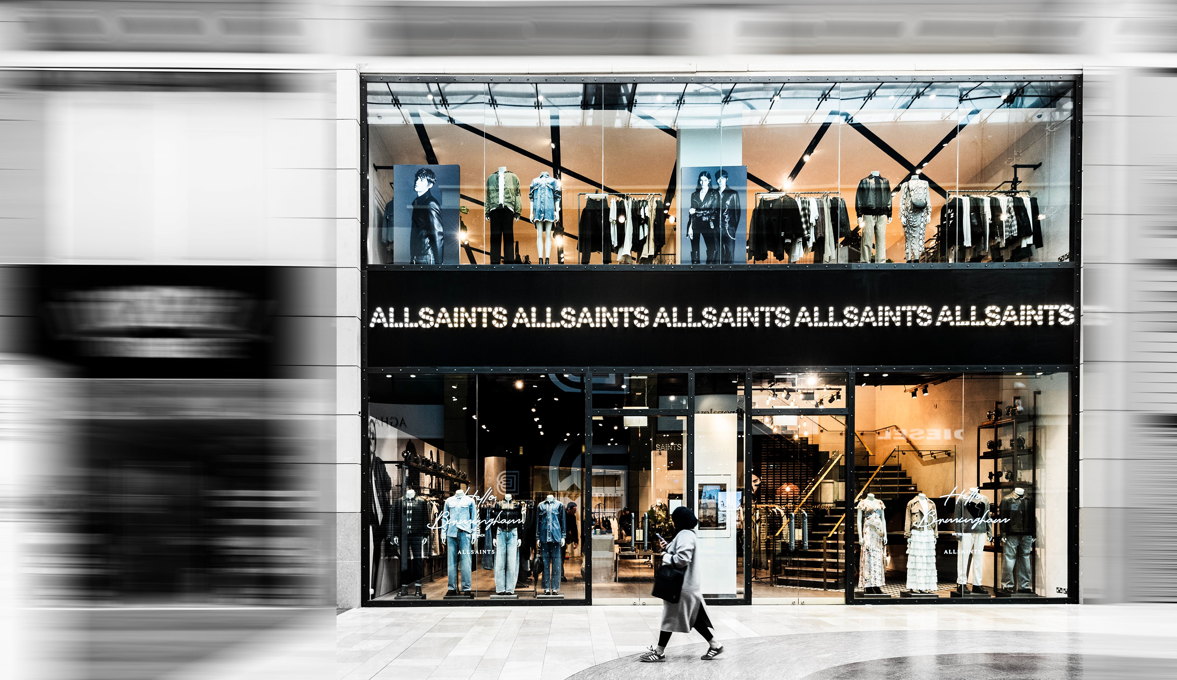 Storefront of an AllSaints fashion store with mannequins displaying trendy outfits. A person walks by, capturing a modern, urban vibe.