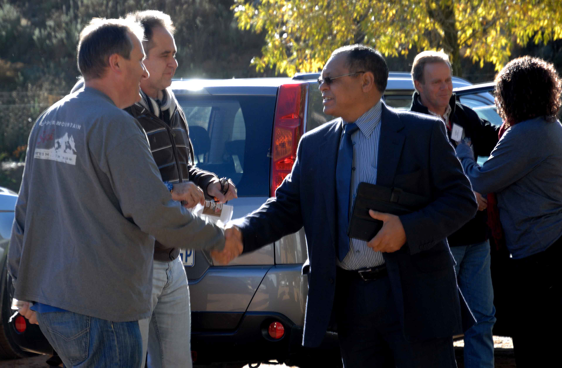 A man in a suit shakes hands with another man in casual clothing outside near parked cars. The scene is sunny, suggesting a friendly, outdoor meeting.