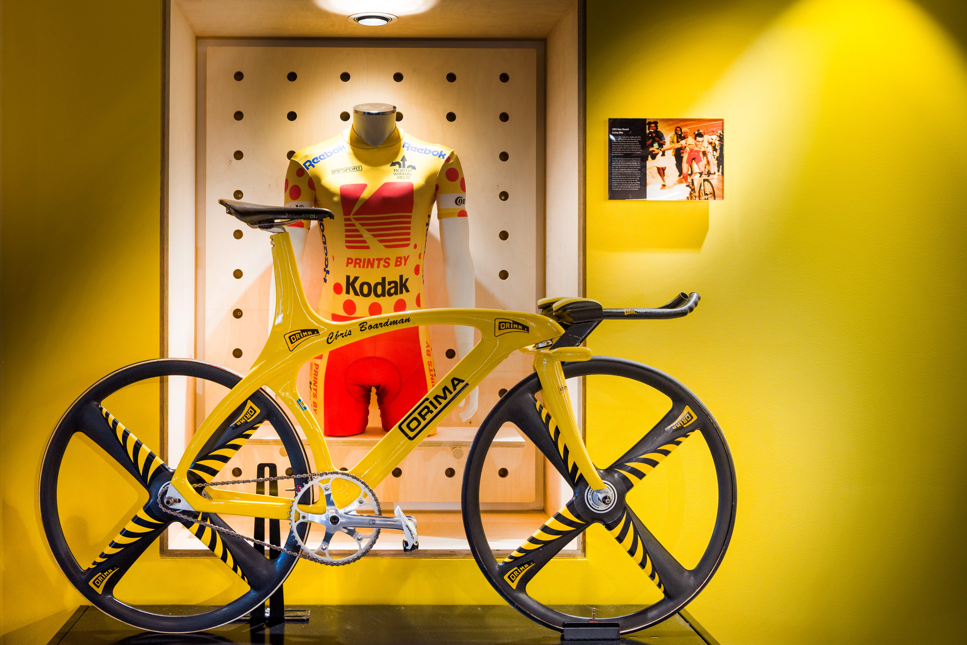 A yellow racing bike with black accents displayed against a yellow wall, next to a matching yellow and red cycling suit. The setup conveys speed and innovation.