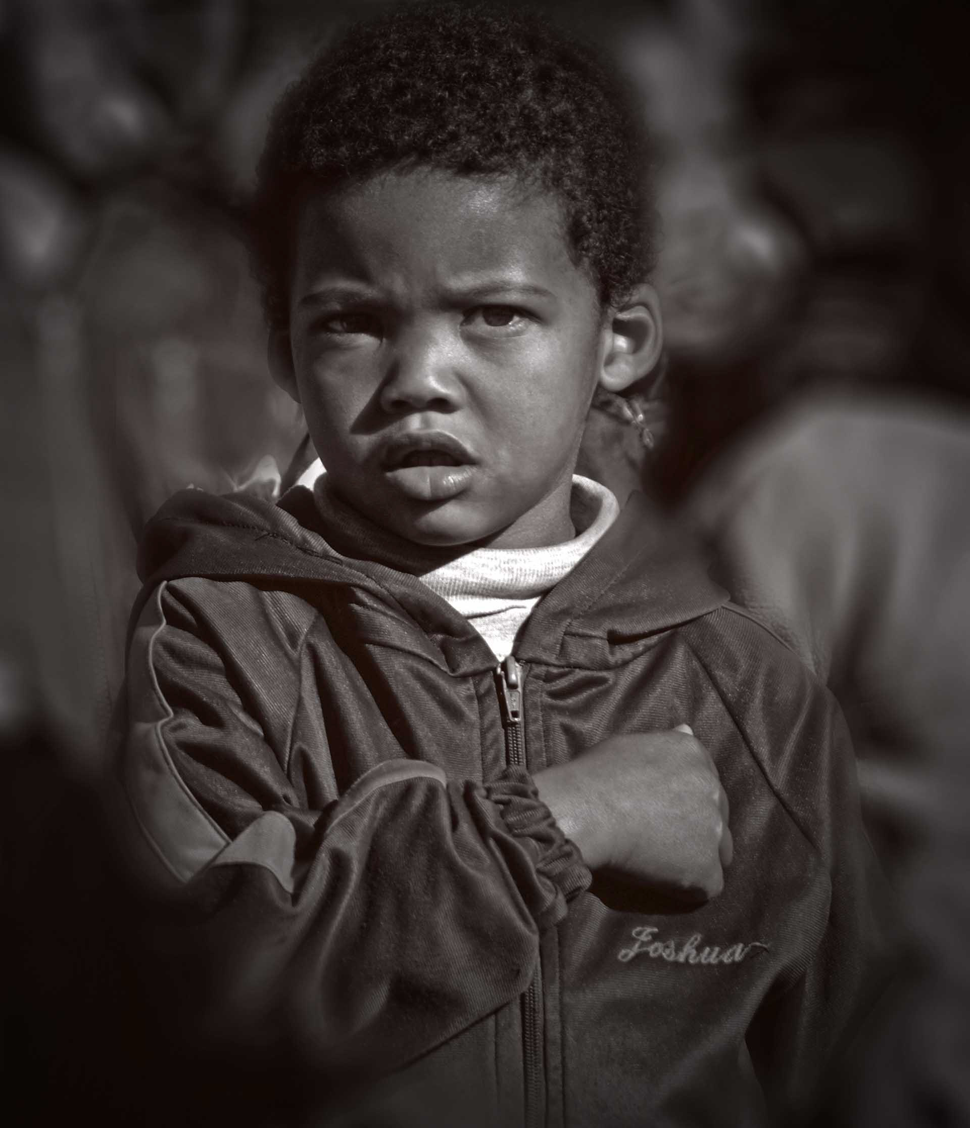 A young child in a hooded jacket looks intently ahead with a serious expression. Their hand is placed over their chest, conveying a solemn tone.