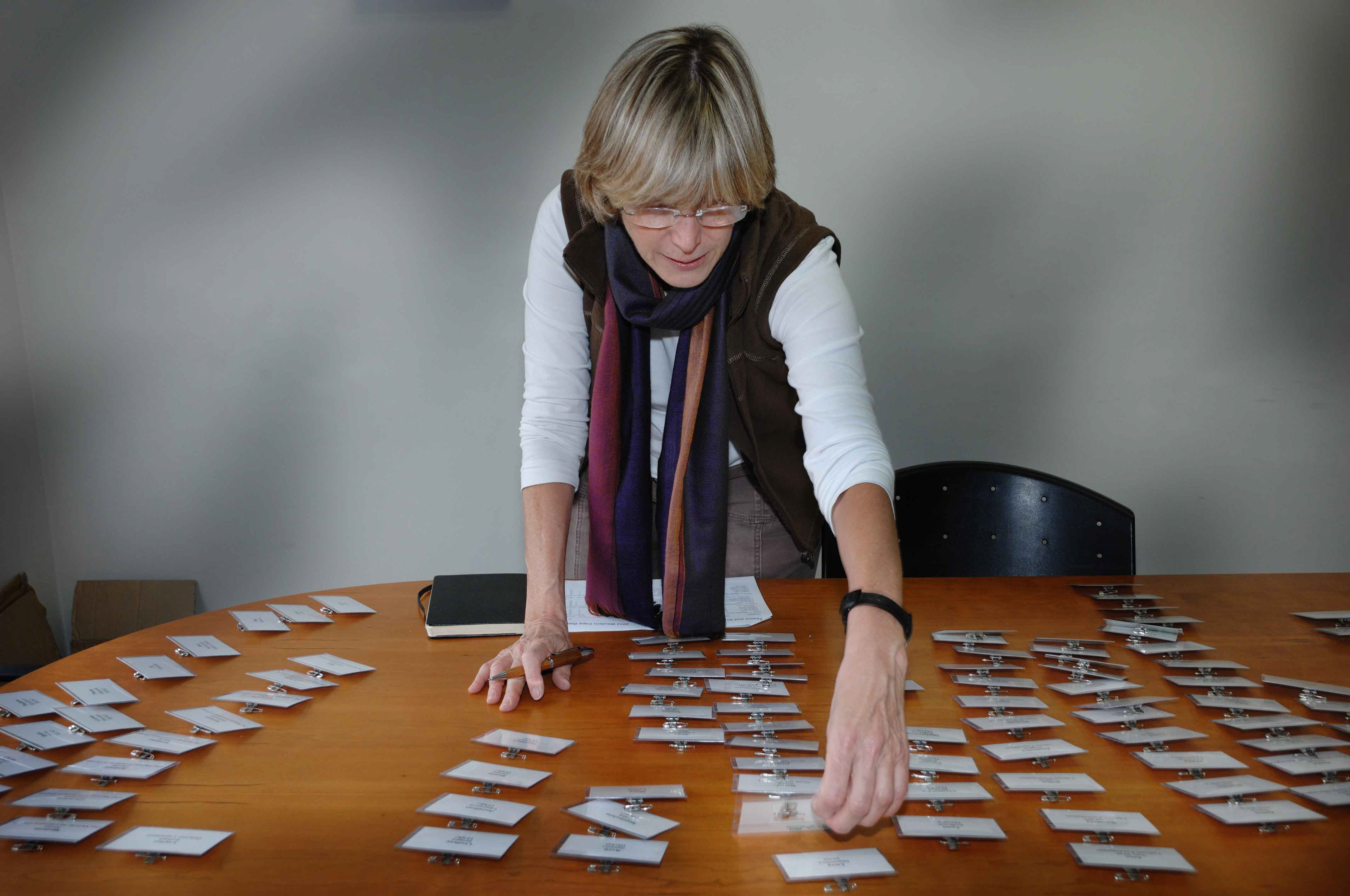 A woman leaning over a wooden table organizes name tags. She's focused, with a notepad nearby, exuding a sense of preparation and orderliness.