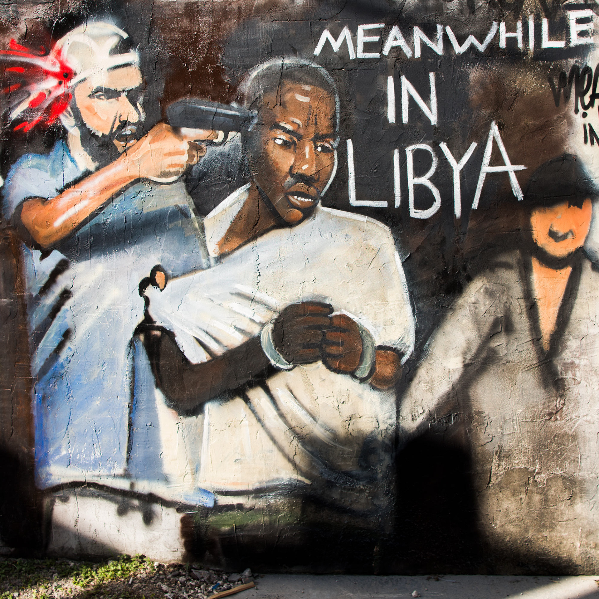 Street art mural shows a man holding a gun to another man's head, who appears frightened. The words "Meanwhile in Libya" are painted beside them.