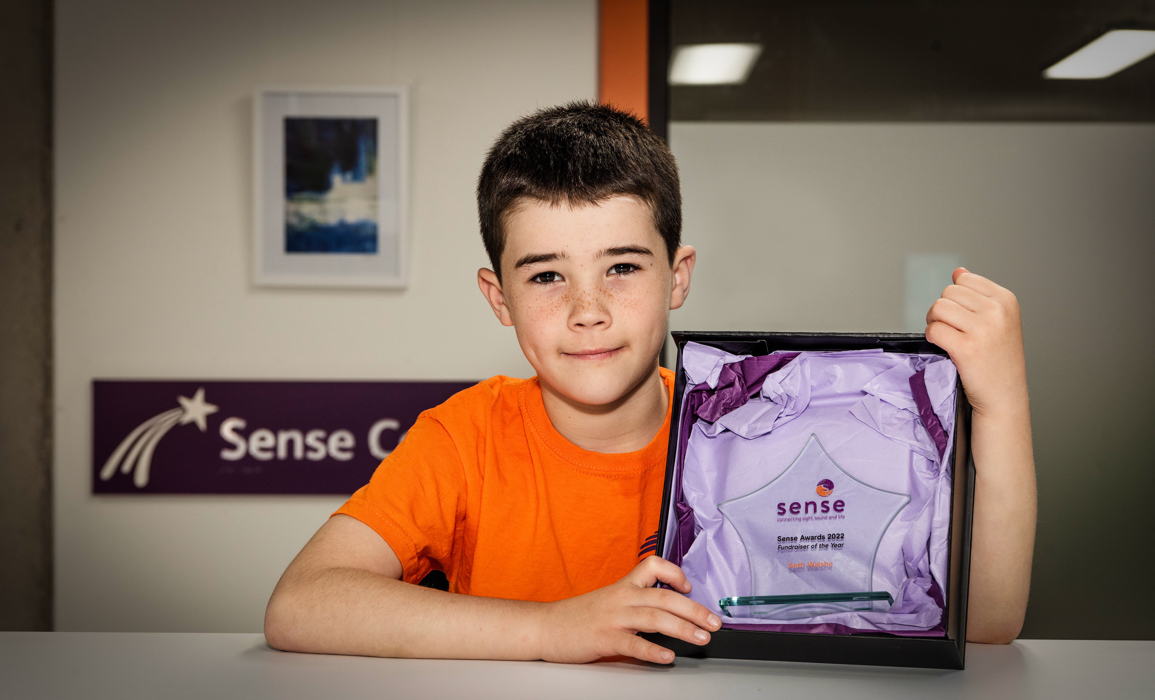 Young boy in an orange shirt proudly holds a star-shaped award, wrapped in purple paper, in front of a "Sense Center" sign. The mood is celebratory. charity photography project professional photography services