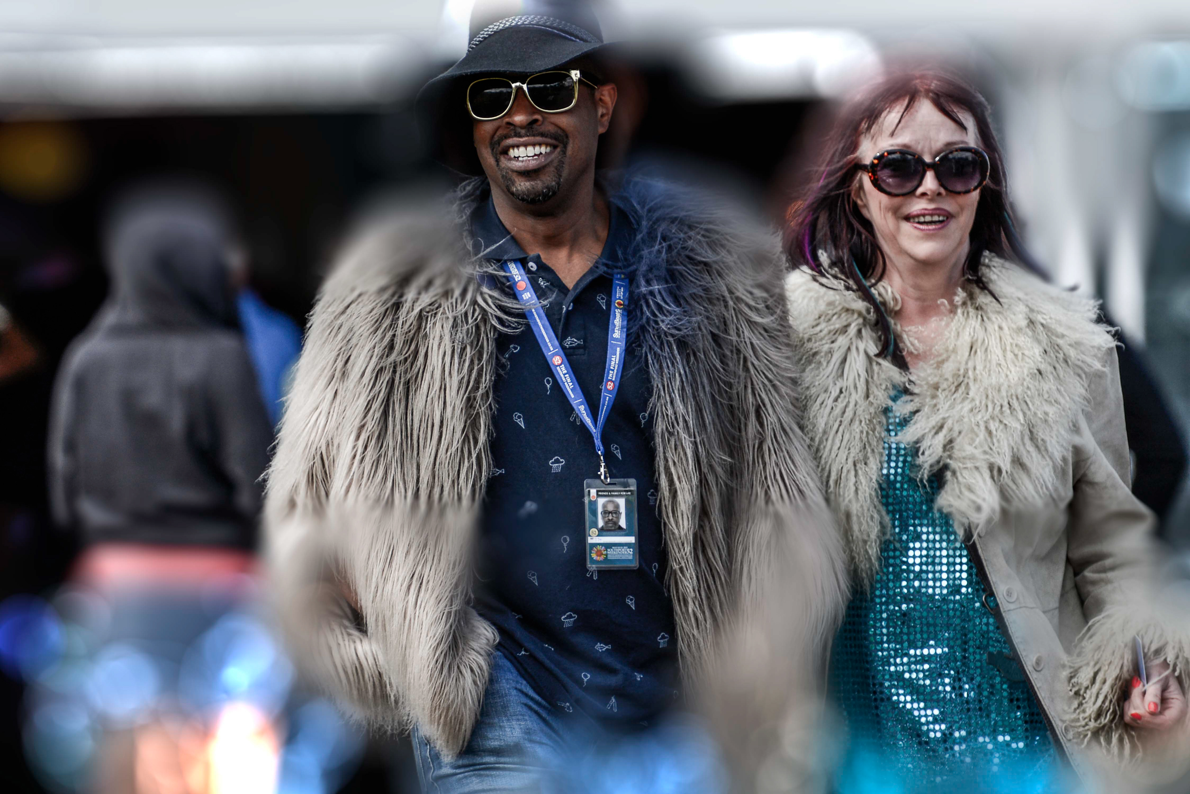 Man and woman in fashionable attire with fur coats and sunglasses, smiling confidently. The man wears a black hat, adding a stylish touch.