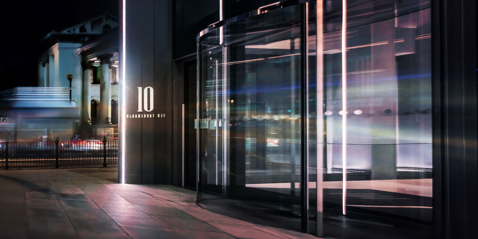 Nighttime street scene with a modern building entrance displaying "10 Bloomsbury Way." Blurred motion of passing cars and lights creates a dynamic, urban atmosphere.