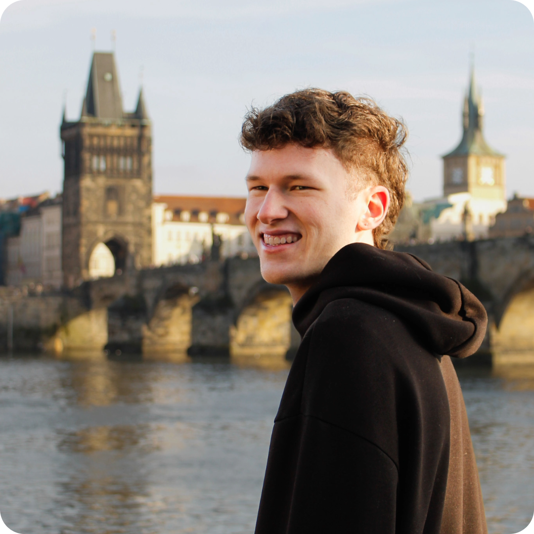 Vincent Ludewig steht am Ufer eines Flusses, im Hintergrund sind historische Gebäude mit Türmen und eine Brücke zu sehen. Er trägt einen schwarzen Kapuzenpullover. Das Licht wirkt warm und weich.