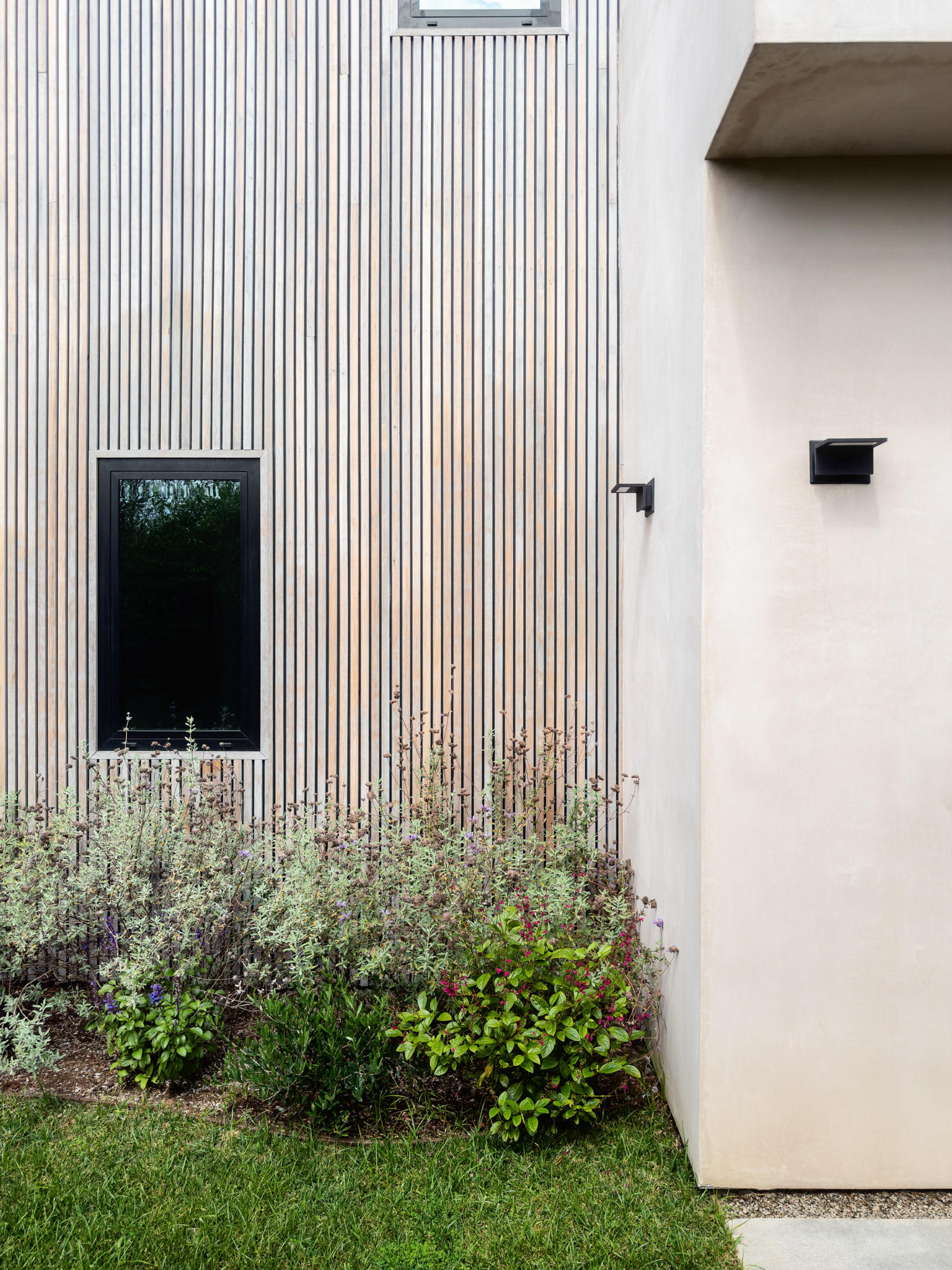 Architectural detail of a modern residential exterior featuring vertical wood siding, smooth stucco, black-framed windows, and landscaped planting.