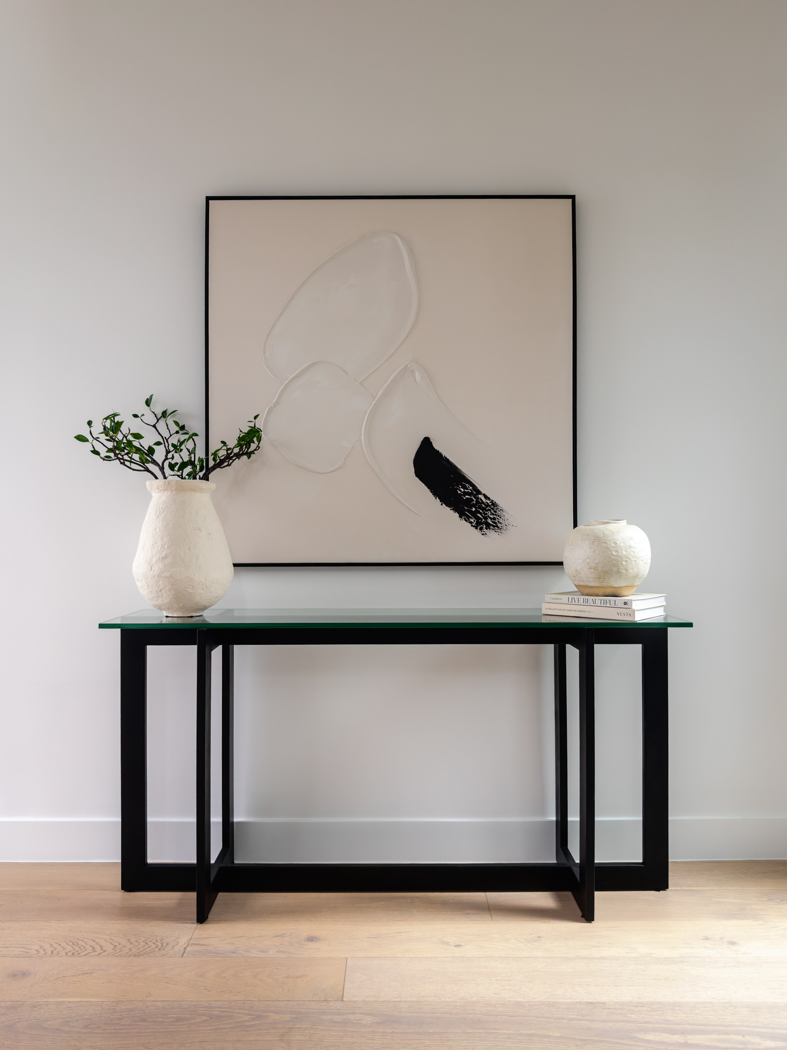 Minimalist hallway interior featuring a black console table, large abstract artwork, sculptural ceramic vases, and warm wood flooring.