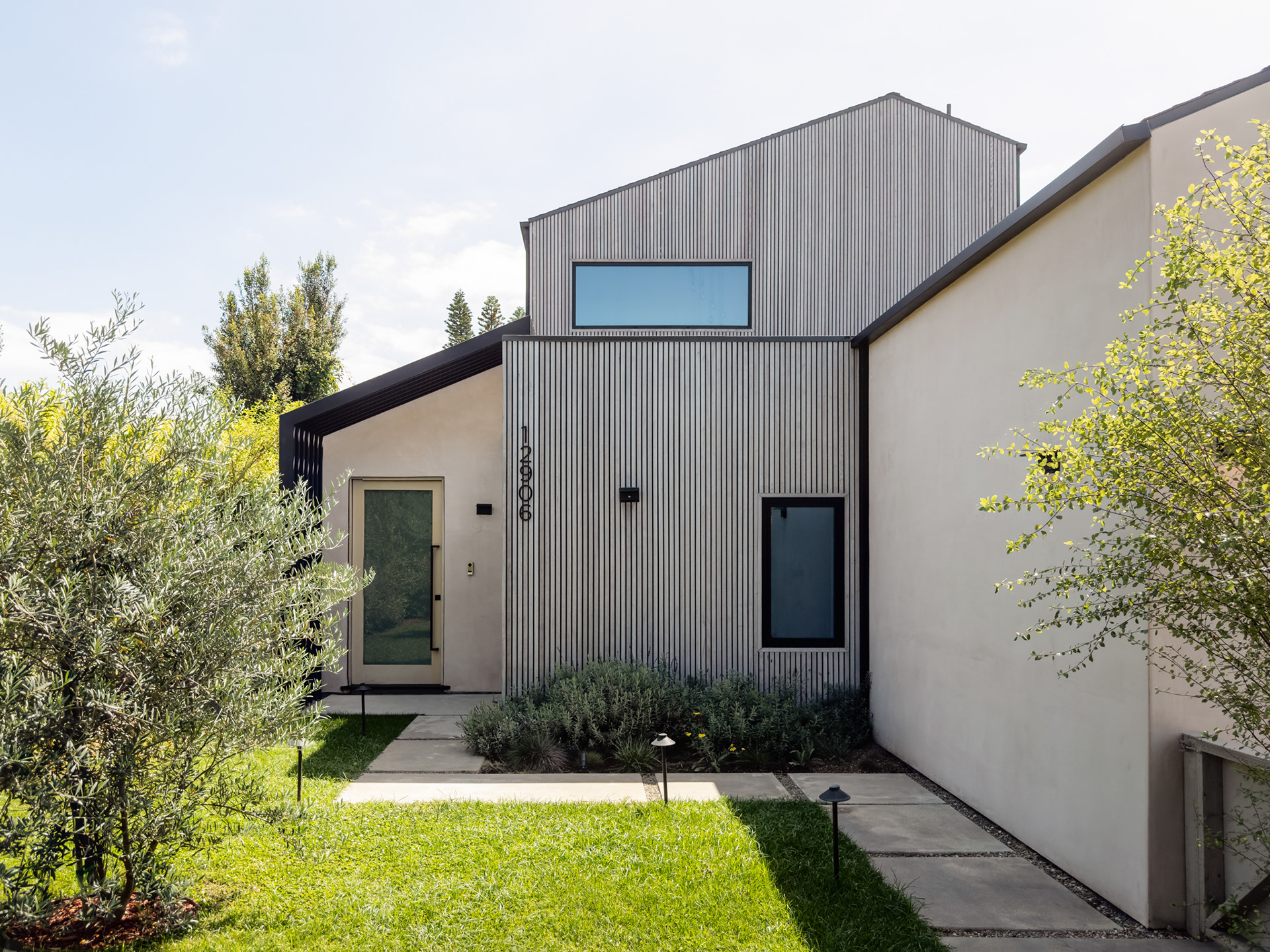 Modern residential exterior featuring vertical wood cladding, smooth stucco walls, black-framed windows, and minimalist landscaping.