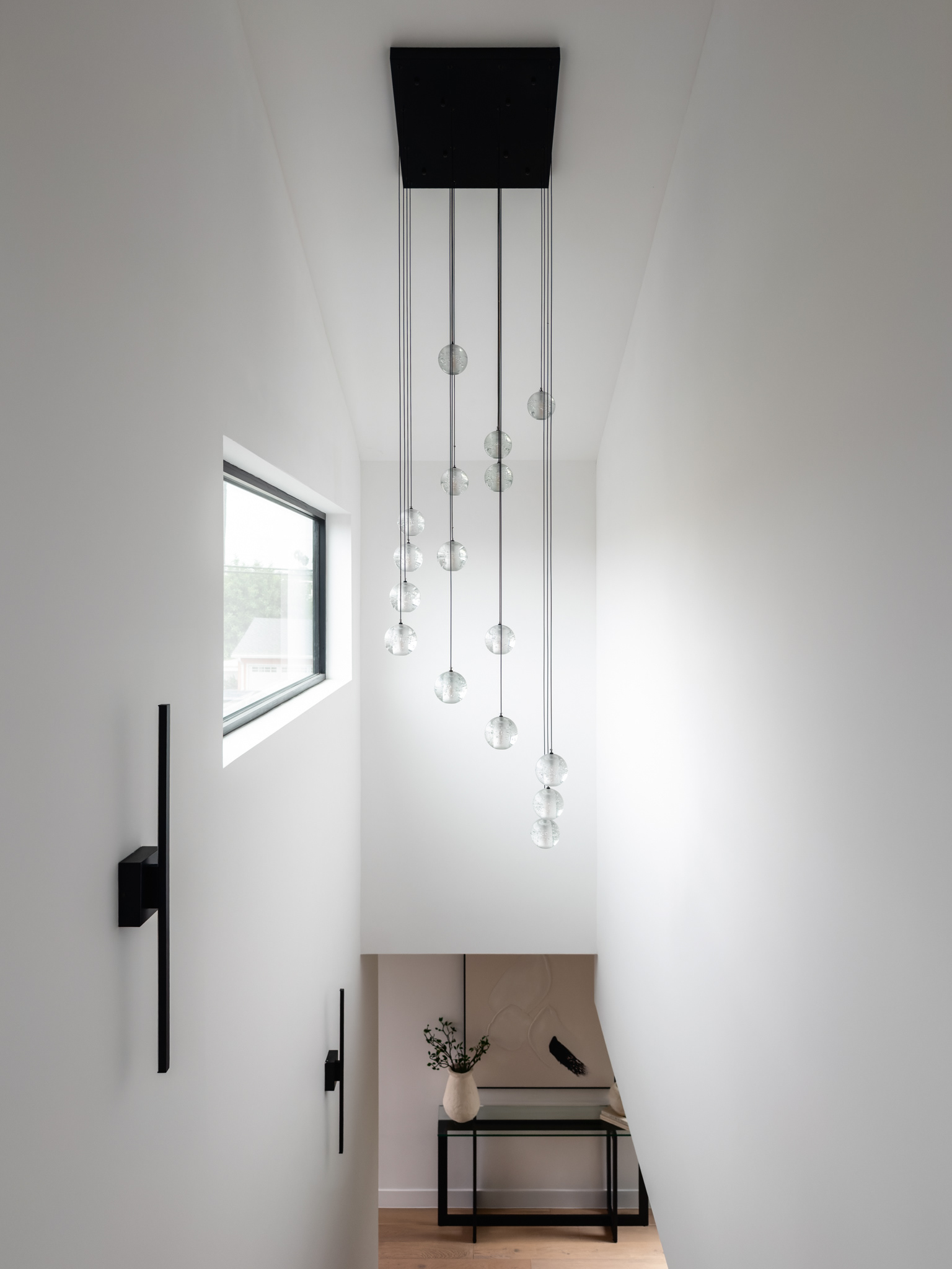 Modern stairwell featuring a cascading glass globe pendant light, minimalist black wall sconces, white walls, and warm wood flooring.
