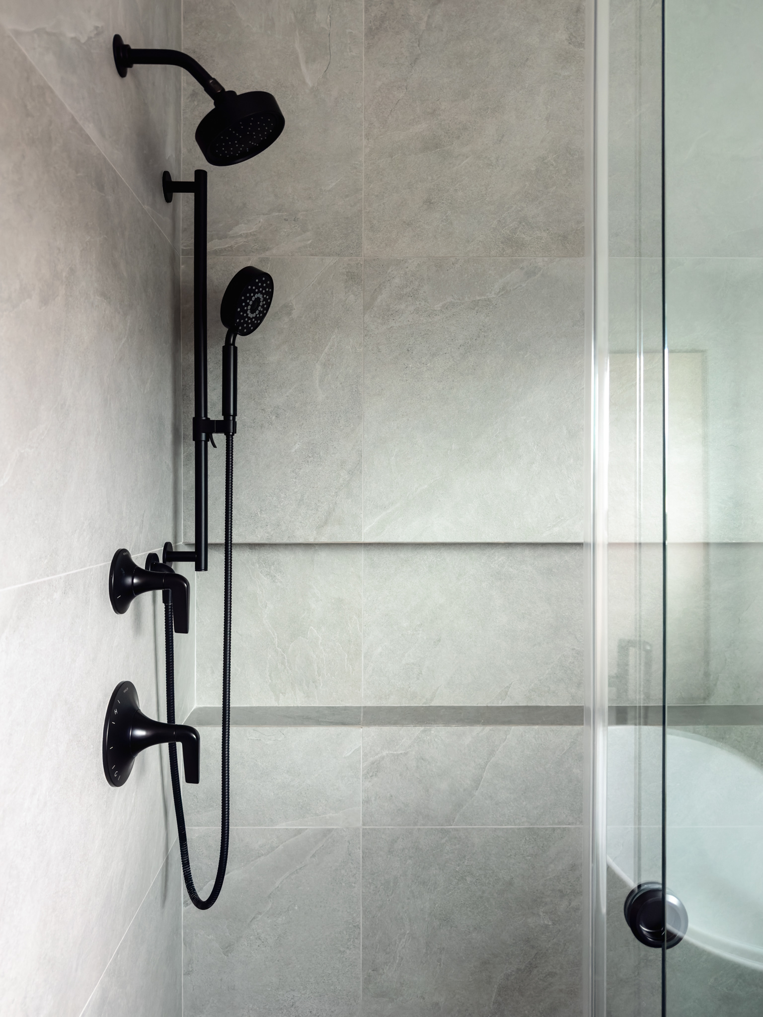 Modern walk-in shower featuring large-format gray tile, matte black rainfall and handheld shower fixtures, a recessed niche, and frameless glass.