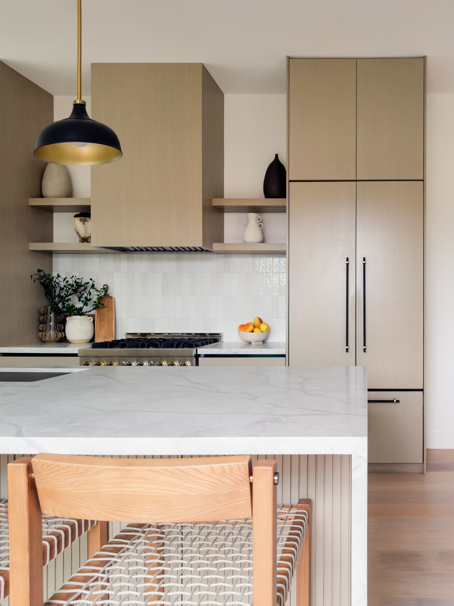 Modern kitchen featuring light wood cabinetry, a marble waterfall island, open shelving, black and brass pendant lighting, and woven counter stools.