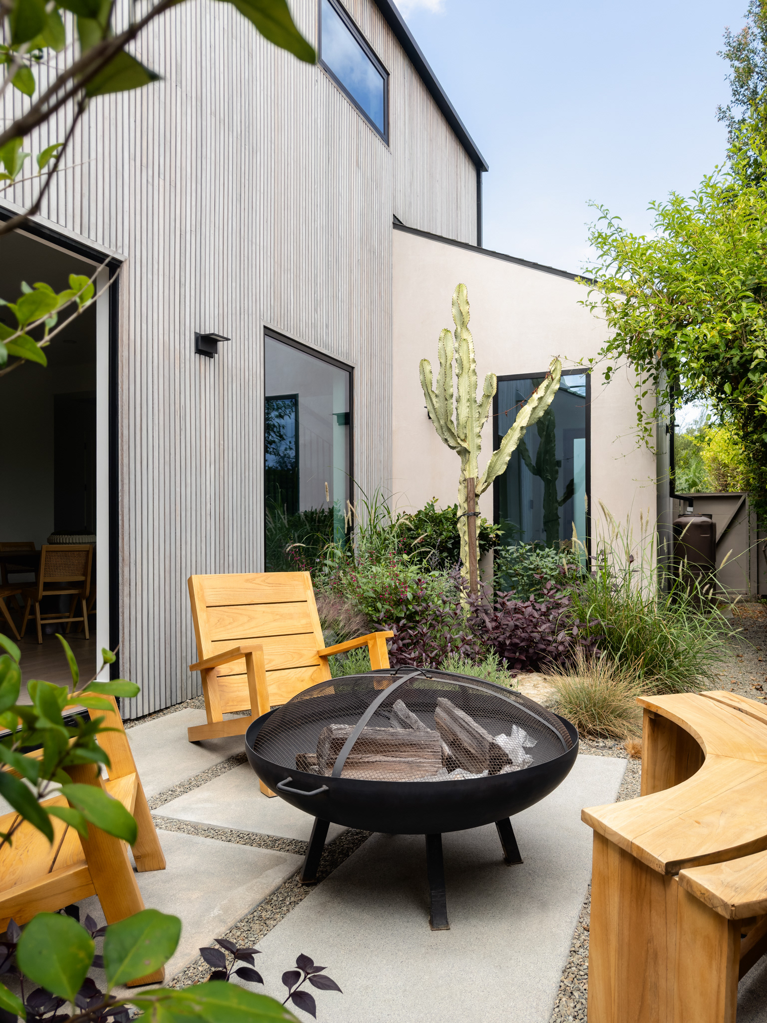 Modern outdoor patio with wood seating, a circular fire pit, lush landscaping, and contemporary vertical wood siding.