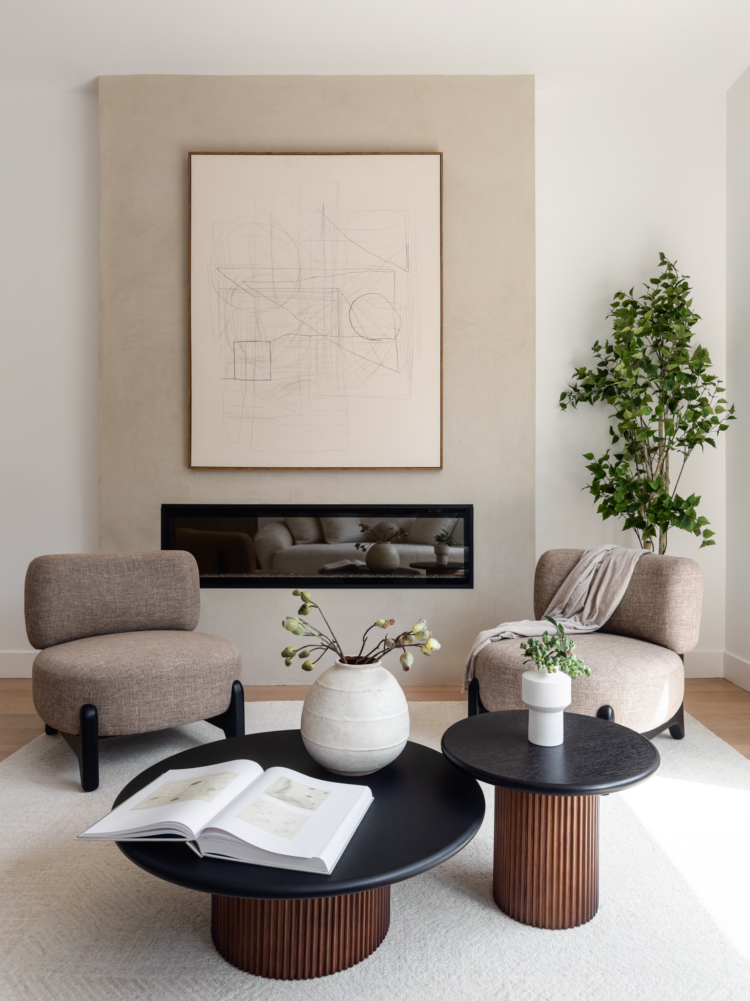 Modern living room featuring sculptural lounge chairs, layered black coffee tables, a minimalist fireplace, abstract artwork, and warm neutral finishes.