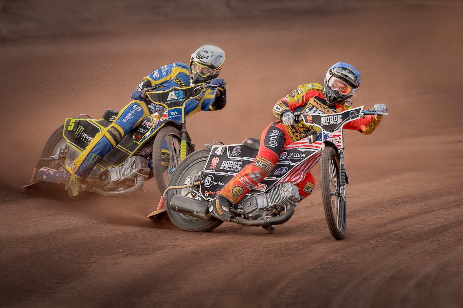 Two speedway racing motorcyclists racing on a dirt track, with the rider in the foreground wearing red and yellow gear and the rider behind in blue and yellow gear. Both riders are leaning into the turn, kicking up dust as they speed around the track. The intense competition and high-speed action are captured in the moment.