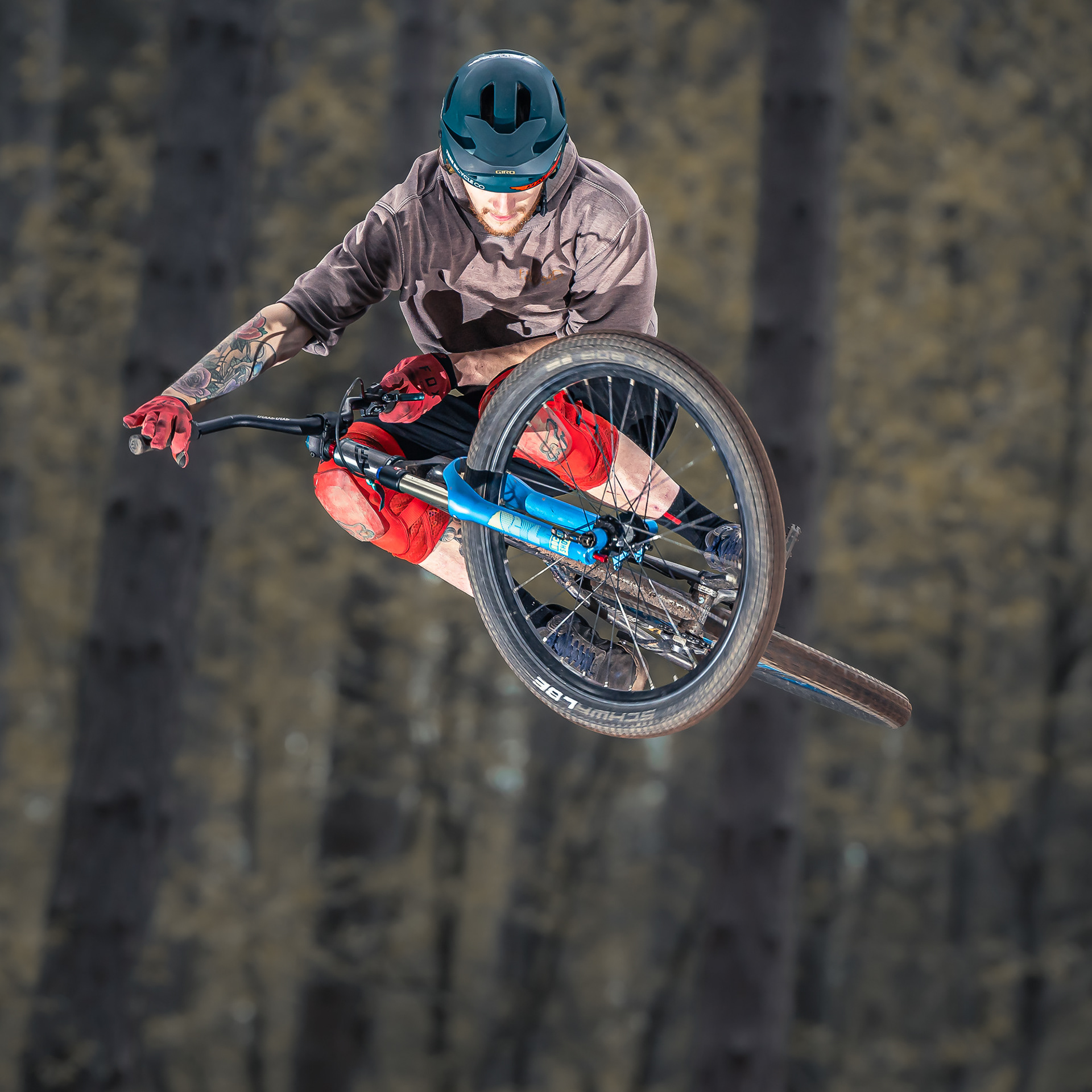 Mountain biker performing an aerial stunt in the forest, wearing a helmet and protective gear. The rider is captured mid-air with the bike tilted sideways, showcasing the skill and excitement of the jump against a backdrop of tall trees.