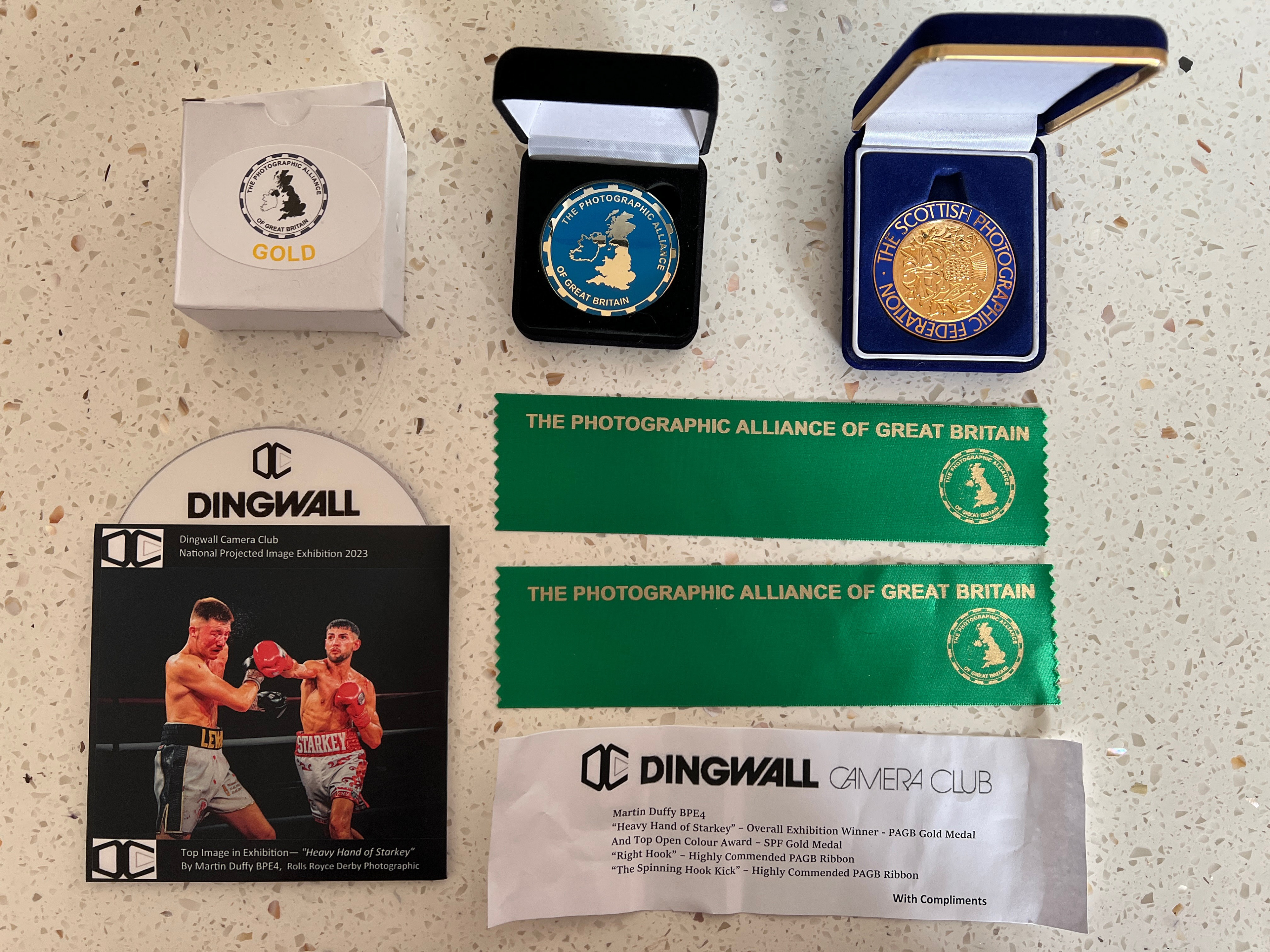 A collection of awards and recognitions displayed on a speckled surface. The items include:  A white box labeled "Gold" with a logo of The Photographic Alliance of Great Britain. Two medal cases: one with a blue and white emblem and the text "The Photographic Alliance of Great Britain" and another with a gold medal and the text "The Scottish Photographic Federation." Two green ribbons with gold text and emblems, stating "The Photographic Alliance of Great Britain." A certificate from Dingwall Camera Club featuring a photo of two boxers in a match, titled "Top Image in Exhibition - 'Heavy Hand of Starkey' By Martin Duffy BPE4, Rolls Royce Derby Photographic." A small paper note from Dingwall Camera Club listing awards: Martin Duffy BPE4 for "Heavy Hand of Starkey" - Overall Exhibition Winner, PAGB Gold Medal, Top Open Colour Award, SPF Gold Medal. "Right Spinning Hook Kick" - Highly Commended PAGB Ribbon. The note ends with "With Compliments."