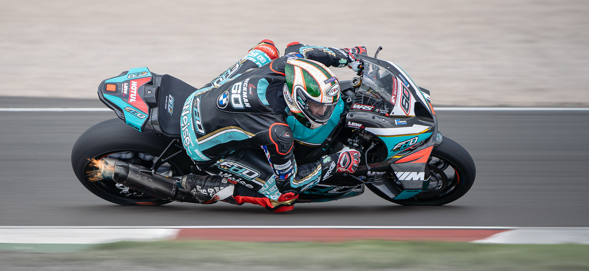 Motorcycle racer in green and black gear, with bike number 60, leaning into a turn on a race track with sparks flying from the exhaust shot by a professional motorsport photographer.