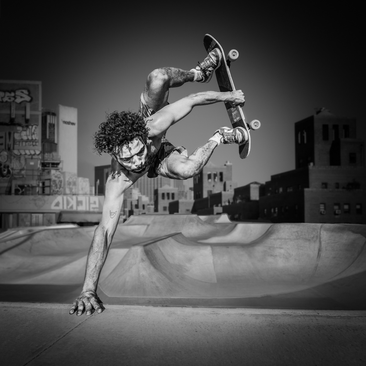 Black and white image of a skateboarder performing an inverted aerial trick in a skatepark, captured mid-air with one hand on the ground and the other holding the skateboard. The background features urban buildings with graffiti, enhancing the gritty, dynamic atmosphere of the scene.