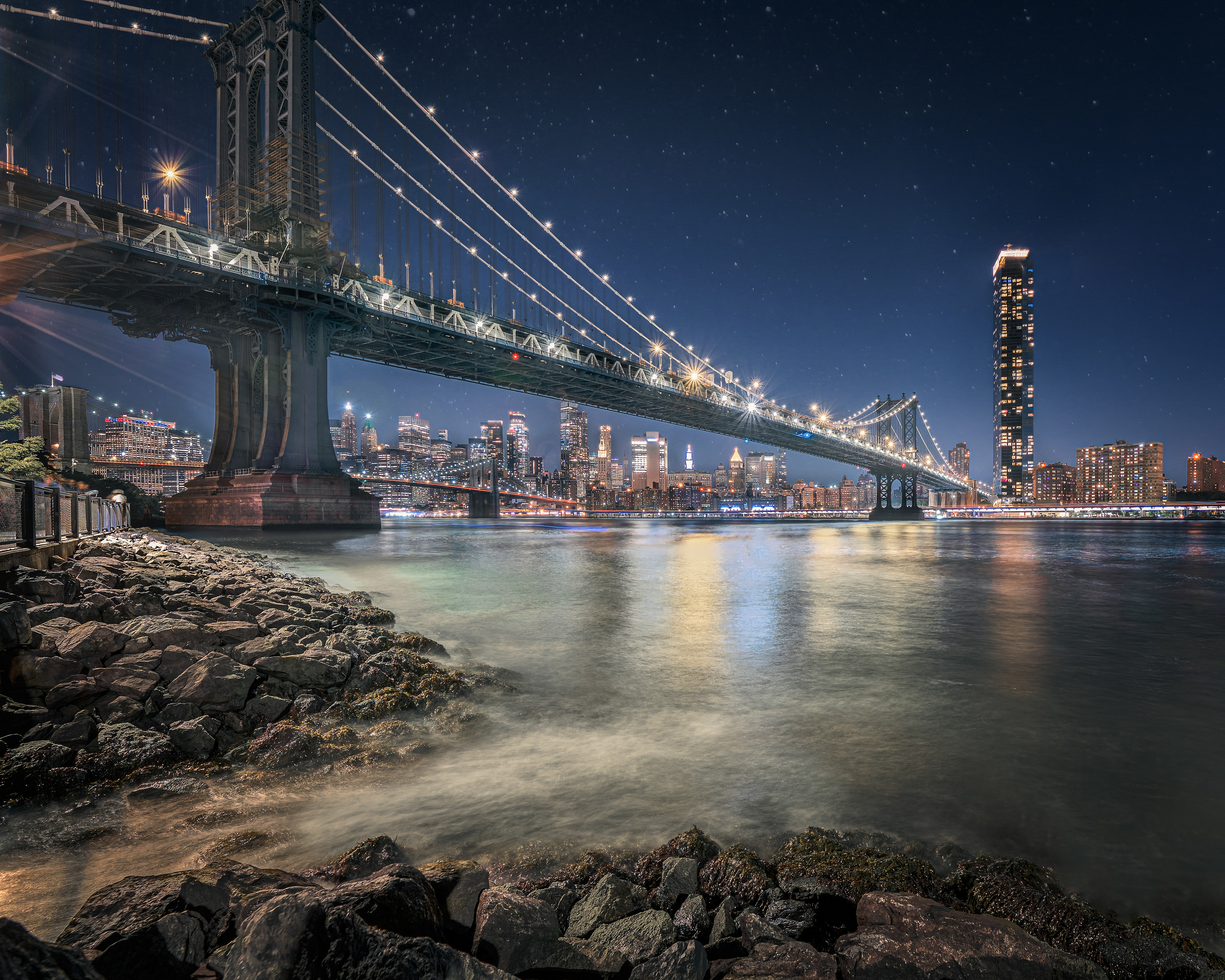 A night view of the Manhattan Bridge spanning across the East River, with the illuminated New York City skyline in the background. The bridge is adorned with lights, and the water below reflects the city lights. The foreground features a rocky shoreline with gentle waves lapping against the stones. The sky is clear, revealing stars above the cityscape.