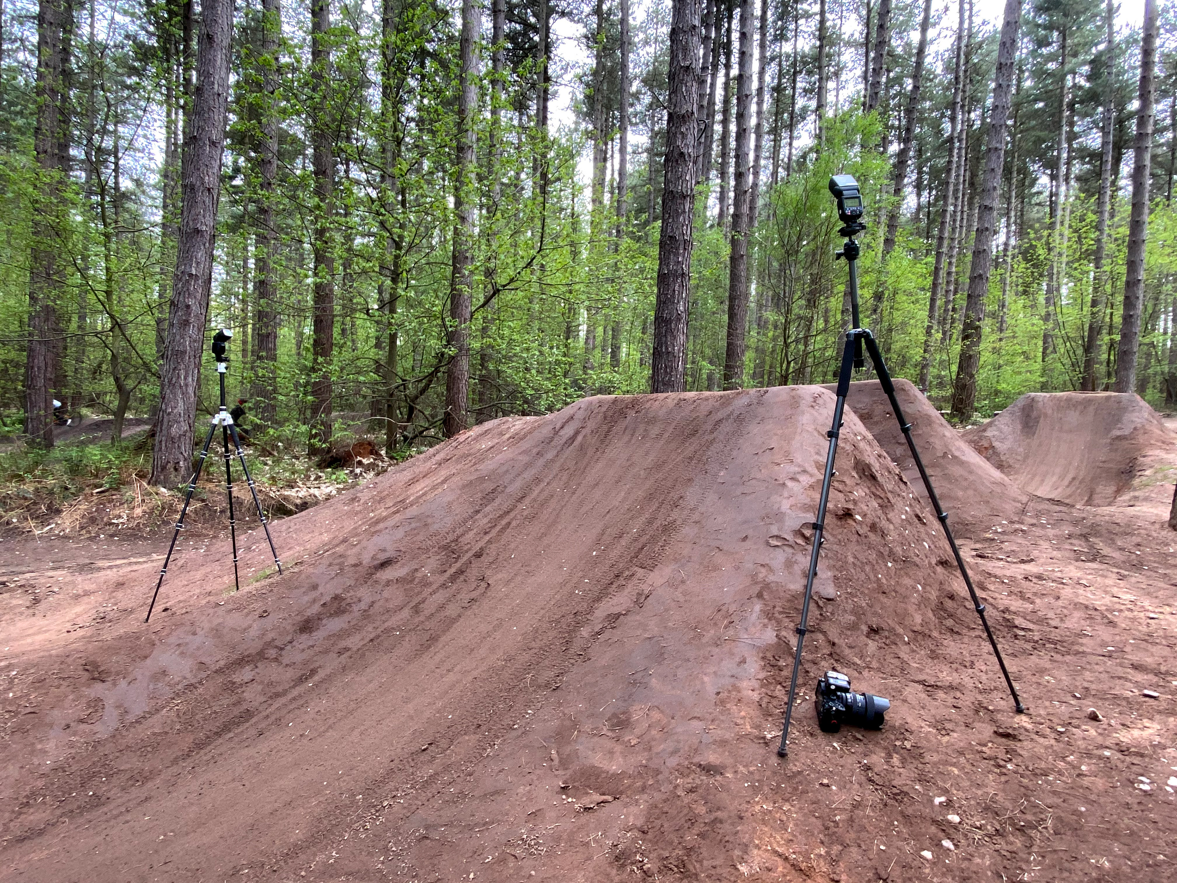 Forest scene with a dirt jump ramp set up for mountain biking, featuring two tripods with cameras and flash units positioned to capture the action. The setting is lush with green trees, indicating a well-prepared spot for photographing bike stunts.
