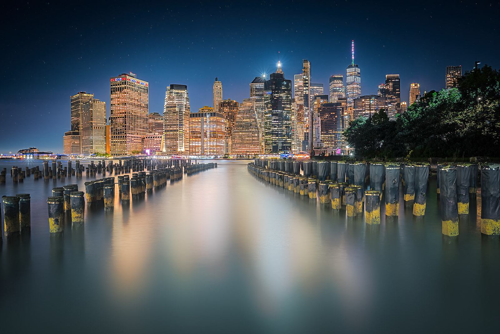 This captivating nighttime shot captures the illuminated New York City skyline, viewed from across the water. The buildings' lights reflect beautifully on the water's surface, creating a vibrant and mesmerizing scene. In the foreground, remnants of old pier pilings stand in neat rows, leading the eye towards the towering skyscrapers. The clear night sky is dotted with stars, adding to the overall serene and majestic atmosphere of this iconic cityscape.