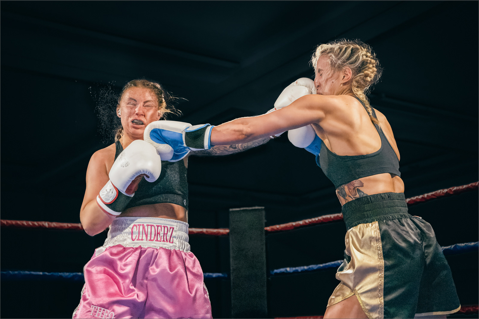 Two female boxers in a ring during a match, with the boxer on the right, wearing black and gold shorts, landing a punch on the boxer on the left, who is wearing pink shorts labeled 'CINDERZ.' The boxer on the left has a pained expression and is recoiling from the impact.