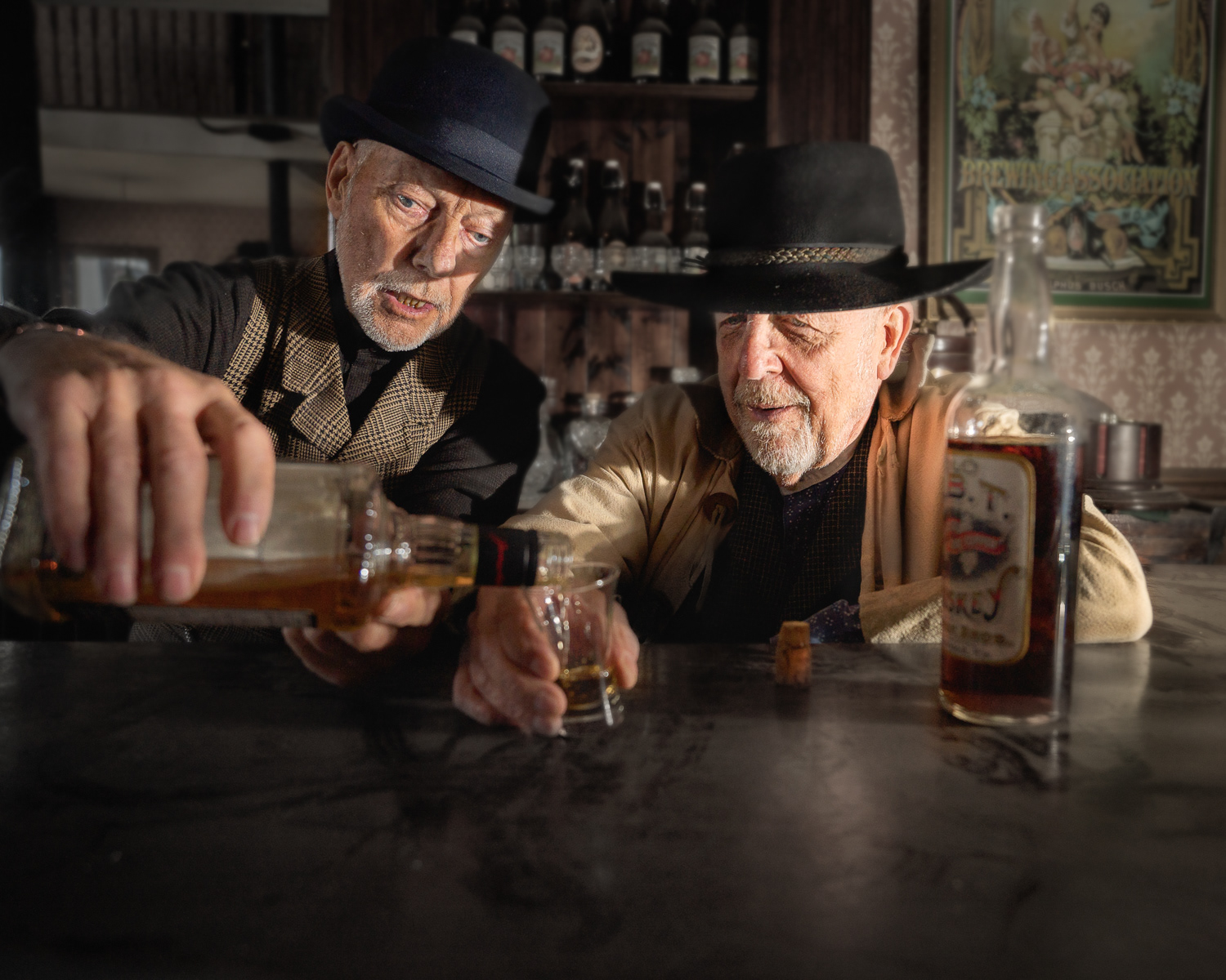 Two elderly men, dressed in vintage attire, are sitting at a rustic bar. One man, wearing a bowler hat and checkered vest, is pouring whiskey from a bottle into a glass for the other man, who is wearing a wide-brimmed hat and a tan jacket. The bar is filled with antique bottles and glassware, and there is a nostalgic poster on the wall in the background. The warm lighting and vintage setting create a cozy, historical atmosphere, reminiscent of an old-time saloon.