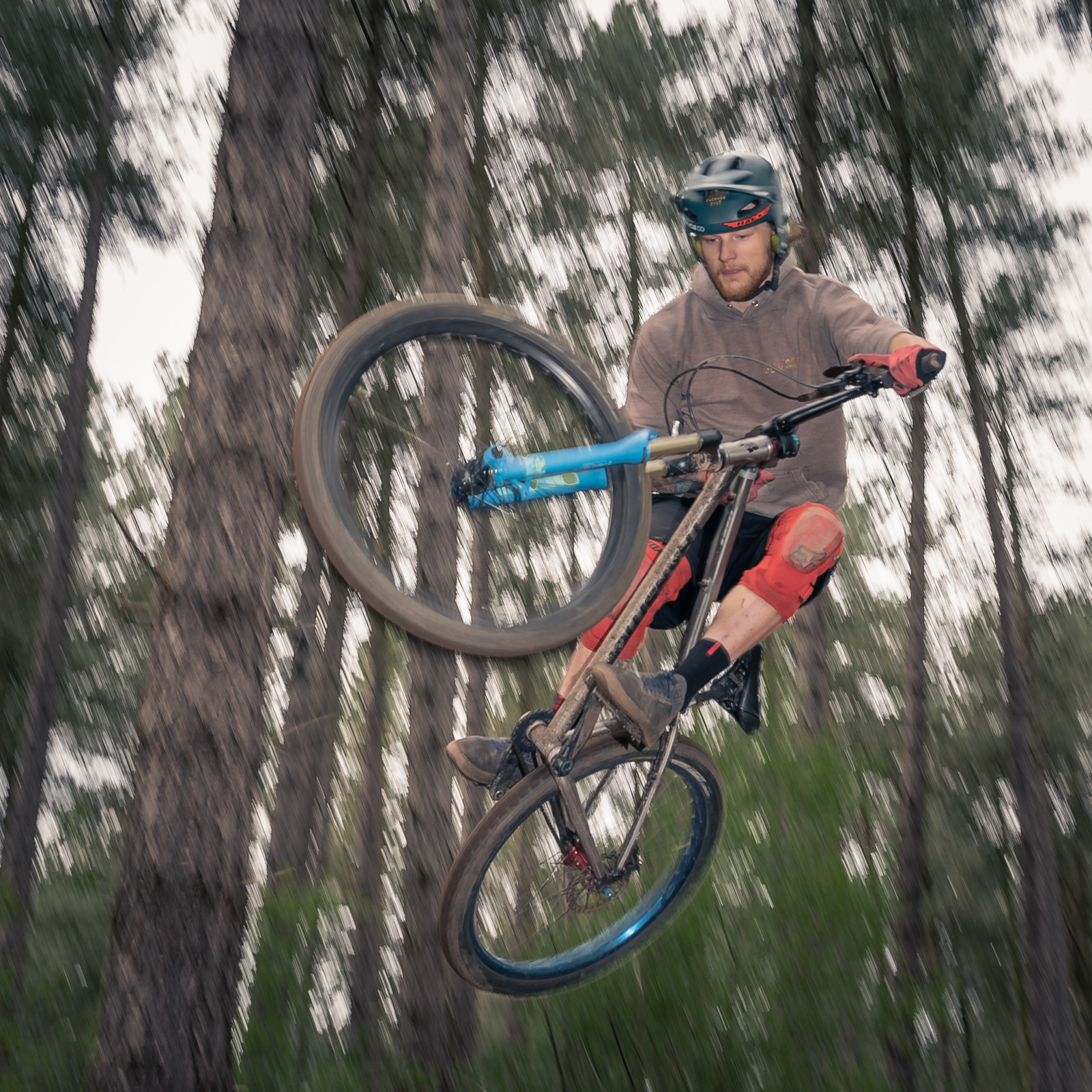 Mountain biker in mid-air performing a jump, wearing a helmet, goggles, gloves, and knee pads. The rider is captured against a backdrop of tall trees in a forest, emphasizing the dynamic and thrilling nature of the sport.