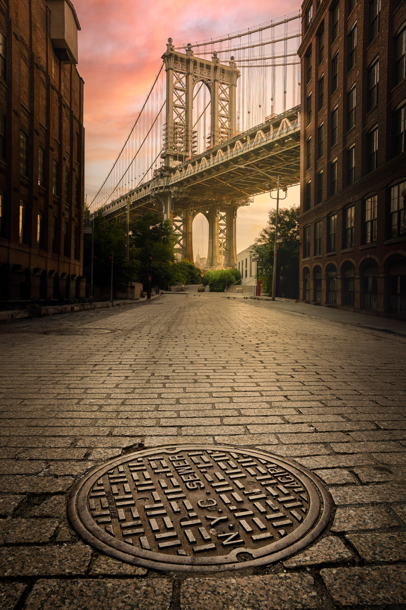 An iconic view of the Manhattan Bridge seen from a cobblestone street in DUMBO, Brooklyn, with the Empire State Building visible through the bridge's supports. The image is captured during a serene sunrise, casting a warm, golden light on the buildings and the street. In the foreground, a circular NYC sewer manhole cover is prominently displayed, adding a touch of urban detail to the scene.