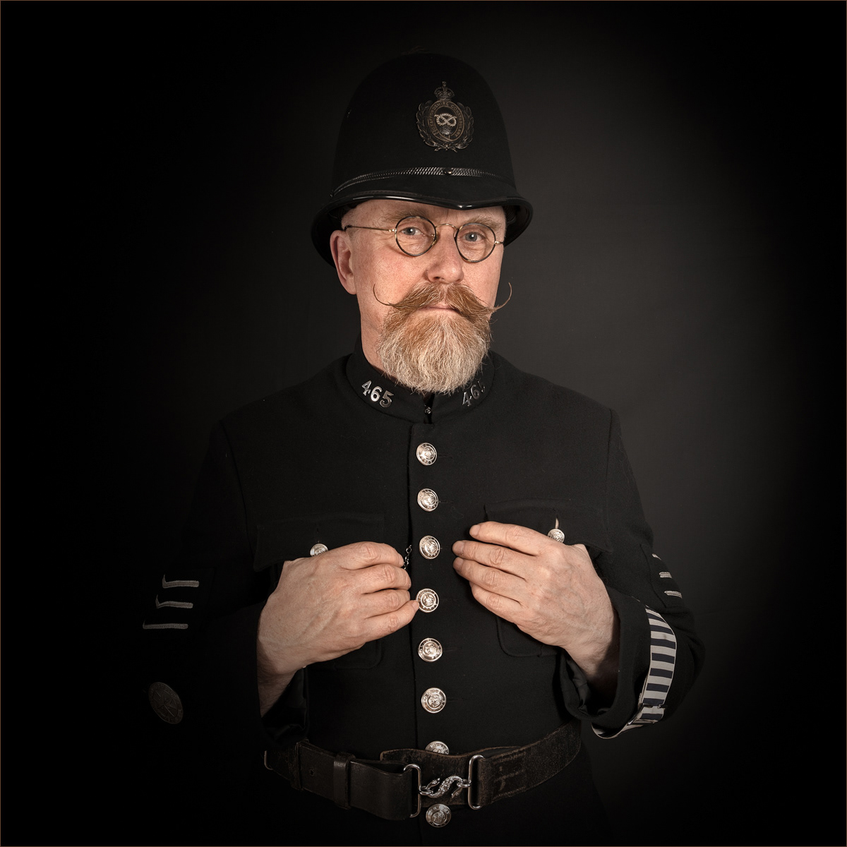 A bearded man dressed in a vintage British police uniform poses against a plain, dark background. He wears a traditional helmet with an insignia, round spectacles, and a dark coat adorned with silver buttons and badges. His hands are adjusting the front of his coat. The man's neatly groomed beard and waxed mustache add to his distinguished and authoritative appearance. The number "465" is visible on his collar, indicating his officer number. The lighting is soft, creating a dramatic and formal portrait.