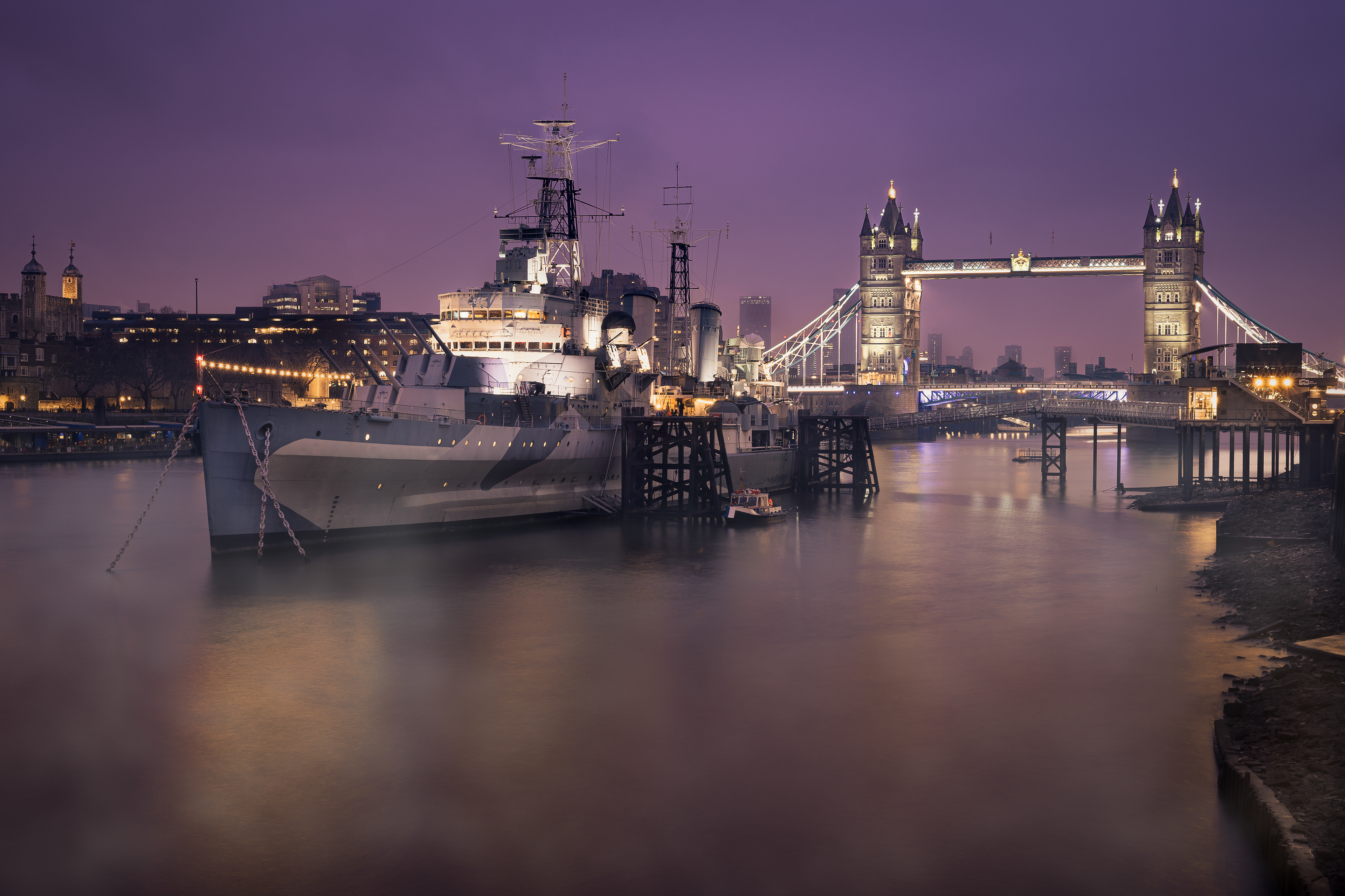A captivating evening view of the iconic Tower Bridge in London, beautifully illuminated against a dusky sky. The HMS Belfast, a historic Royal Navy light cruiser, is prominently anchored in the foreground, its detailed structure and imposing presence adding depth to the scene. The calm waters of the River Thames reflect the lights from the bridge and the surrounding buildings, enhancing the tranquil yet majestic ambiance. The soft purple hue of the sky and the city lights create a harmonious blend, highlighting the historical and architectural grandeur of this London landmark.