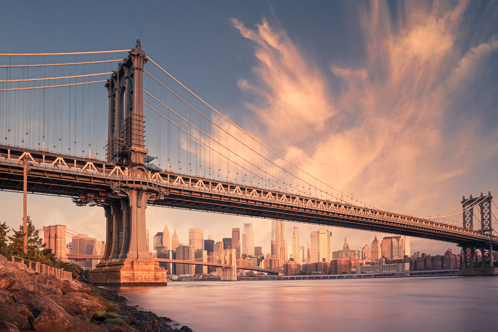 A picturesque view of the Manhattan Bridge in New York City during a vibrant sunset. The bridge spans across the East River with the city skyline in the background, featuring iconic skyscrapers bathed in golden light. The sky is painted with dramatic clouds, enhancing the warm and serene atmosphere of the scene.