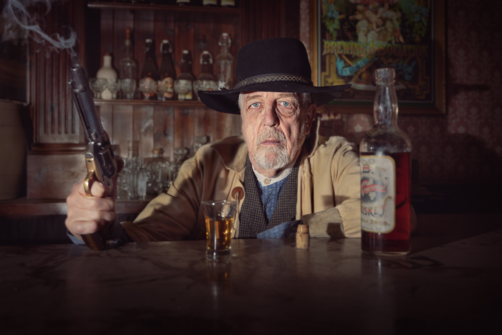 An elderly man dressed as a cowboy from the Old West sits at a bar, holding a smoking revolver in one hand and a glass of whiskey in the other. He is wearing a black wide-brimmed hat, a tan jacket, and a blue shirt with a vest. The background features shelves with various bottles and a vintage poster on the wall, enhancing the historical saloon atmosphere. His expression is stern and serious, conveying the intensity of the moment. The lighting casts dramatic shadows, adding to the gritty, authentic feel of the photograph.