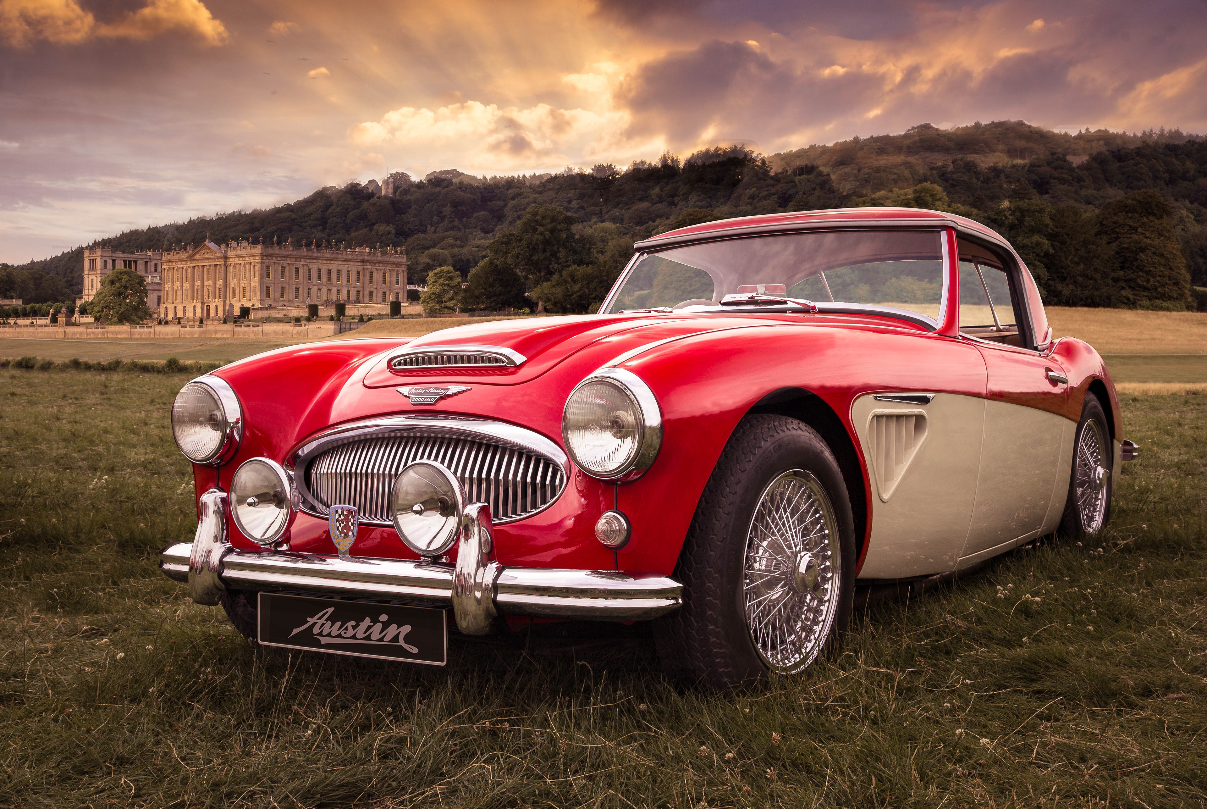 Classic red and white Austin Healey sports car parked on a grassy field with a historic Chatsworth House mansion and wooded hills in the background. The sunset sky casts a warm glow over the scene, highlighting the car's vintage design and chrome details.