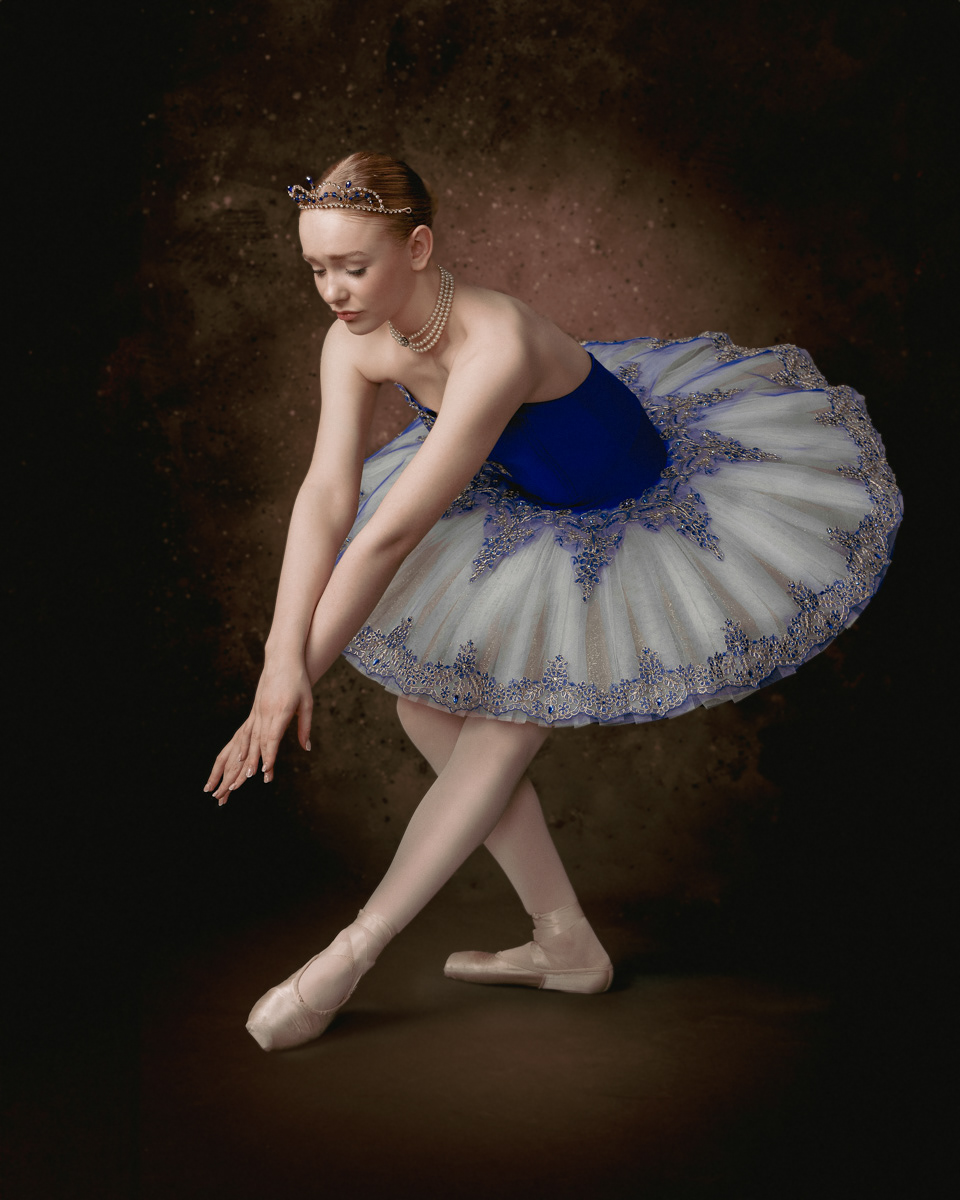 Ballet dancer wearing a blue and white tutu with intricate embroidery, posing gracefully on pointe shoes against a dark, textured background. She is adorned with a tiara and a string of pearls, capturing an elegant and poised moment.