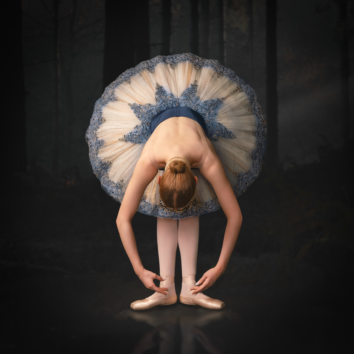 Ballet dancer wearing a blue and white tutu with intricate embroidery, bending forward to adjust her pointe shoes. She stands on a reflective surface against a dark, forested background, capturing a serene and focused moment.