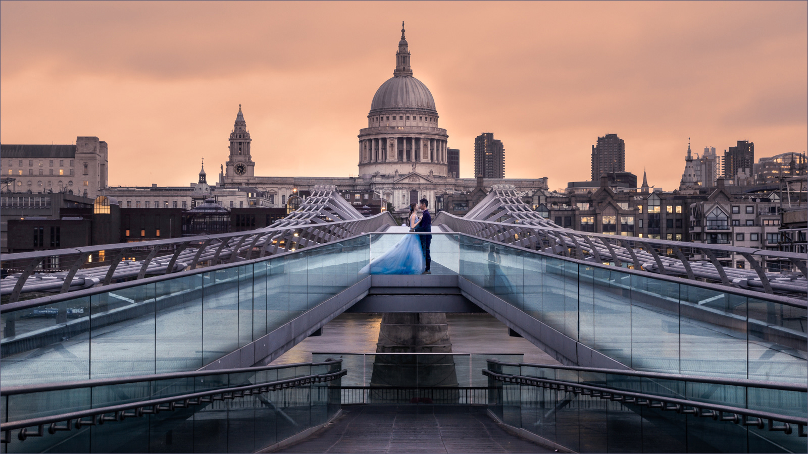 A stunning photograph capturing a couple standing on the Millennium Bridge in London at sunset. The bride wears a flowing blue dress and the groom a dark suit, creating a striking contrast against the modern steel bridge. In the background, the majestic dome of St. Paul's Cathedral is prominently featured, bathed in the warm, golden light of the setting sun, which casts a soft glow over the cityscape. The serene and romantic ambiance is further enhanced by the reflection of the couple and the bridge on the glass panels.