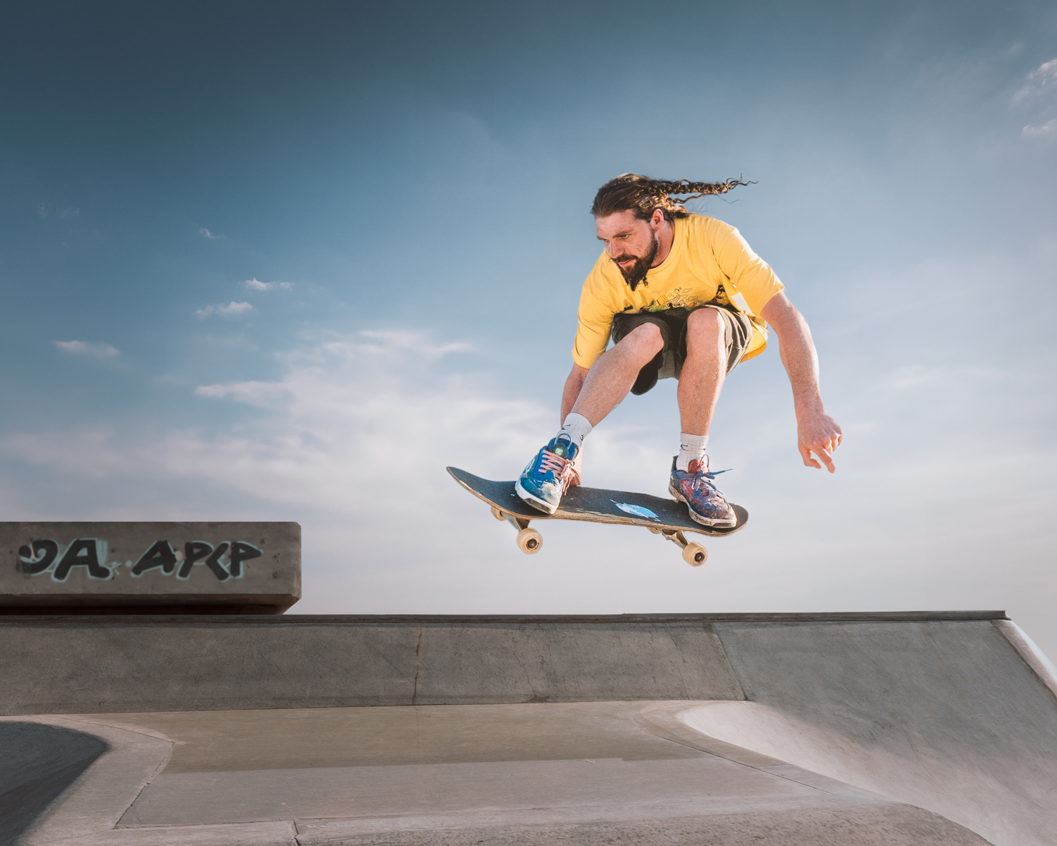 Skateboarder performing an ollie in a skatepark, captured mid-air with knees bent and focused expression. The skater is wearing a yellow shirt, shorts, and colorful sneakers, with long hair flowing behind. The clear blue sky and concrete ramps provide a crisp, clean background for the action.