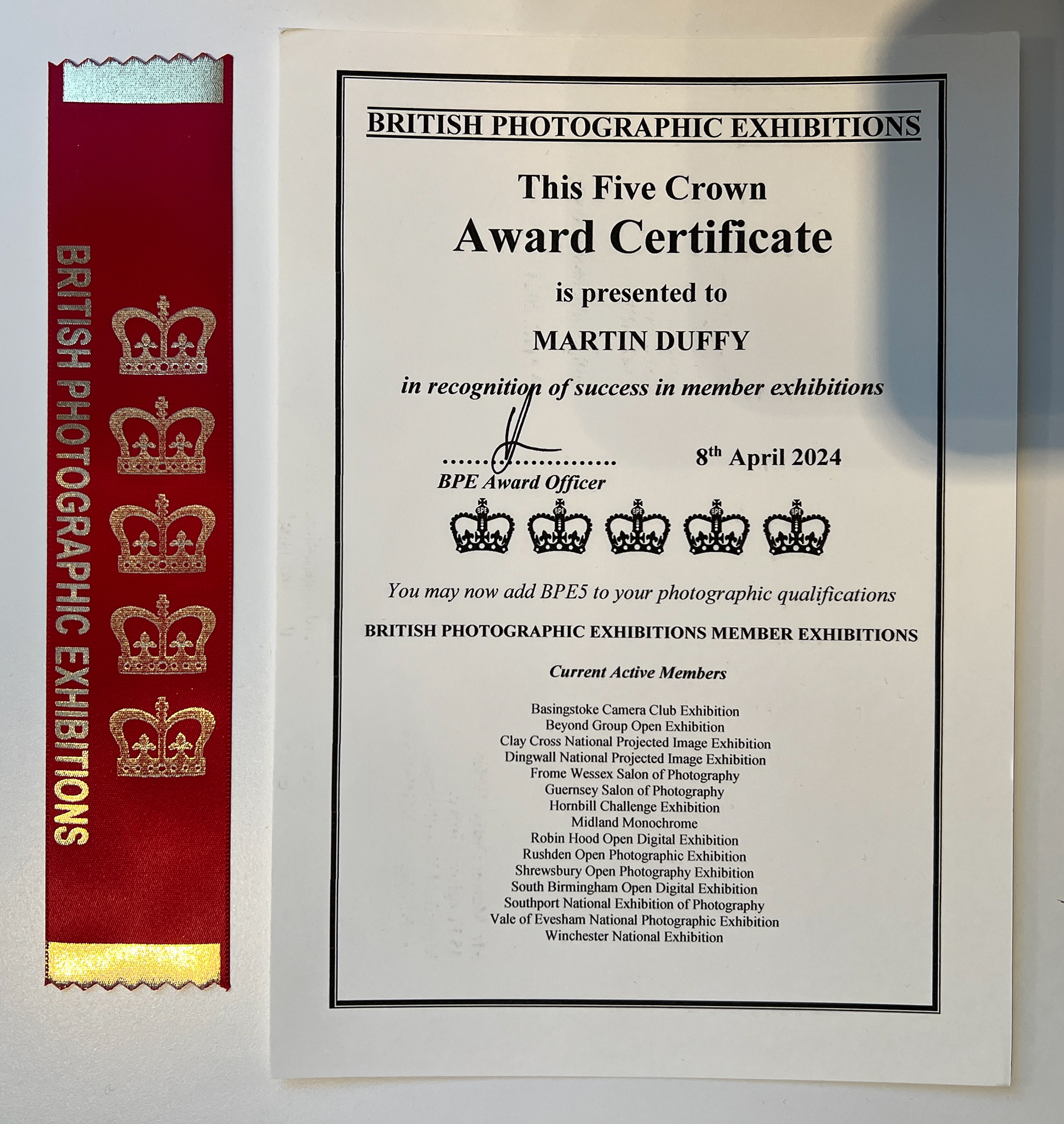 A certificate from the British Photographic Exhibitions (BPE) titled "This Five Crown Award Certificate." The certificate is awarded to Martin Duffy in recognition of success in member exhibitions. It is dated 8th April 2024 and signed by the BPE Award Officer. The certificate includes five crown icons and states that Martin Duffy may now add BPE5 to his photographic qualifications. On the right side of the certificate, there's a list of "Current Active Members" which includes various camera clubs and photographic exhibitions across the UK. A red ribbon with silver text and crown icons is attached on the left side of the certificate, reading "British Photographic Exhibitions."