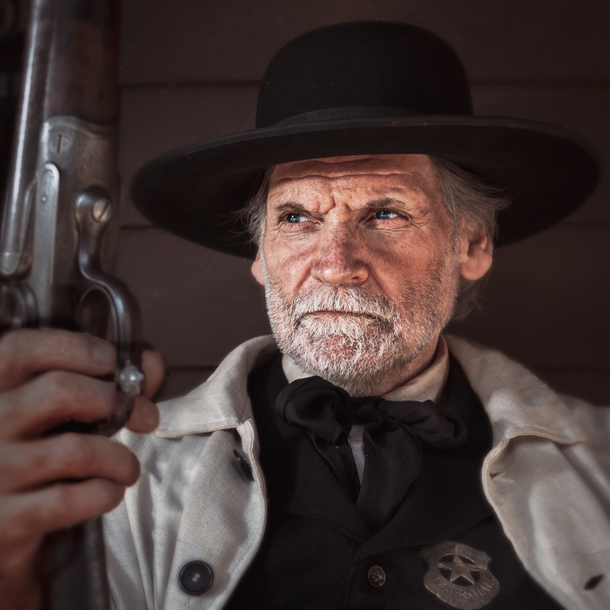 A close-up portrait of an elderly man dressed as a sheriff from the Old West. He wears a black wide-brimmed hat, a light-colored coat, and a black vest with a star-shaped sheriff's badge pinned to it. His face is weathered, and his expression is stern and focused as he holds a rifle upright in one hand. The background is plain, emphasizing the subject's intense and determined demeanor. The lighting highlights the rugged details of his face, adding to the authenticity of the historical portrayal.