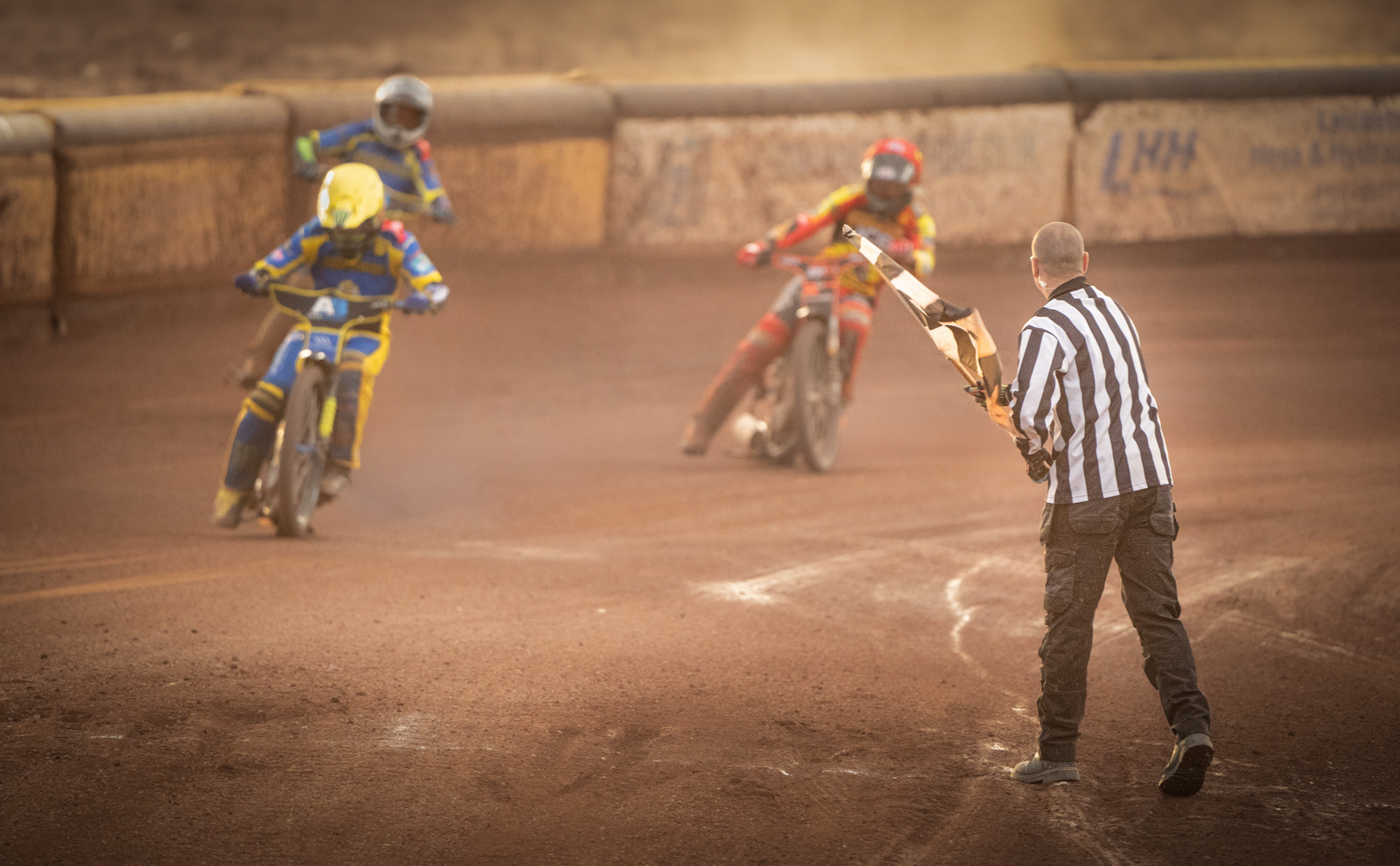 Three speedway motorcyclists racing on a dirt track, with a referee in a striped shirt waving a checkered flag. The leading rider, in yellow and blue gear, is closely followed by another rider in similar gear and a third rider in red and yellow gear. Dust is rising from the track, emphasizing the high-speed action.