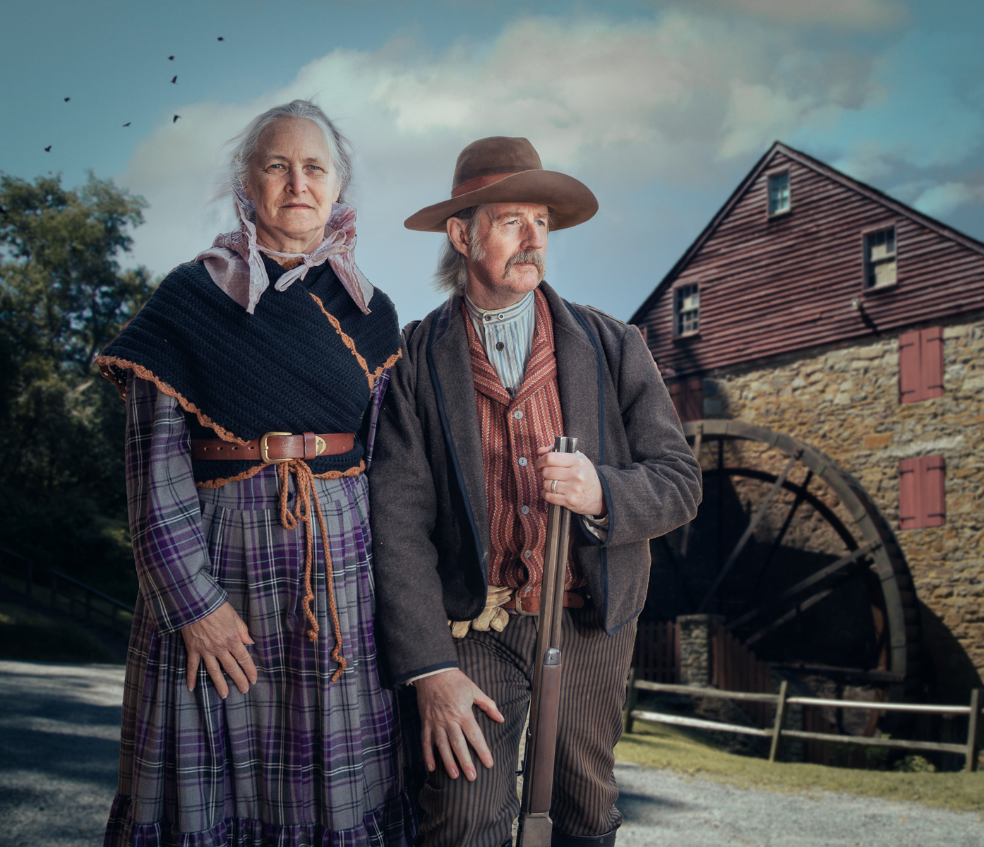 An elderly couple dressed in 19th-century attire stands in front of an old stone building with a large waterwheel. The woman is wearing a plaid dress with a knitted shawl and a bonnet, while the man, with a mustache and a hat, is holding a shotgun and wearing a long coat over striped pants and a vest. The background includes trees and a clear sky, giving the scene a historical and rustic feel. Their expressions convey a sense of pride and resilience.