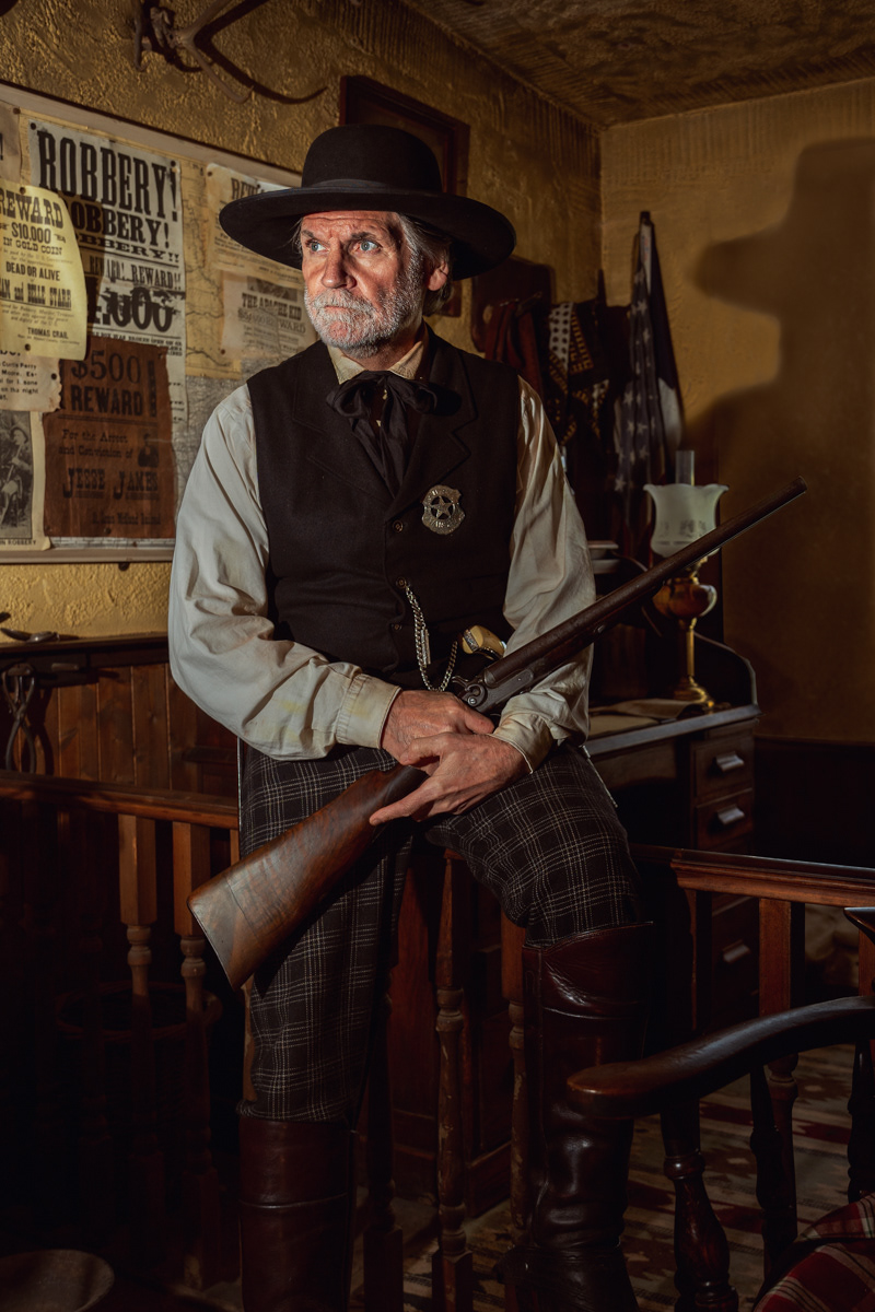 An elderly man dressed as a sheriff from the Old West stands in an office, holding a shotgun. He is wearing a black wide-brimmed hat, a vest with a sheriff's badge, a white shirt, and checkered pants tucked into tall leather boots. Behind him, the wall is adorned with wanted posters and various western decor items. His expression is serious and focused, adding to the historical and law enforcement atmosphere of the scene. The lighting casts a dramatic shadow of his hat on the wall, enhancing the vintage feel of the photograph.