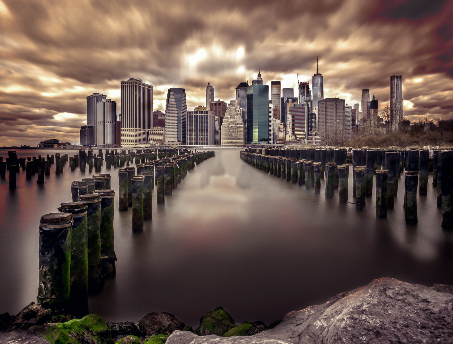 A dramatic view of the Manhattan skyline from Brooklyn, with the decaying wooden pilings of an old pier leading the eye towards the towering skyscrapers. The sky is filled with thick, swirling clouds, casting an ominous yet captivating glow over the cityscape. The long exposure effect creates a smooth, almost ethereal appearance on the water, contrasting with the gritty texture of the moss-covered pilings in the foreground. The overall composition captures the juxtaposition of nature and urban development, highlighting the dynamic and ever-changing character of New York City.