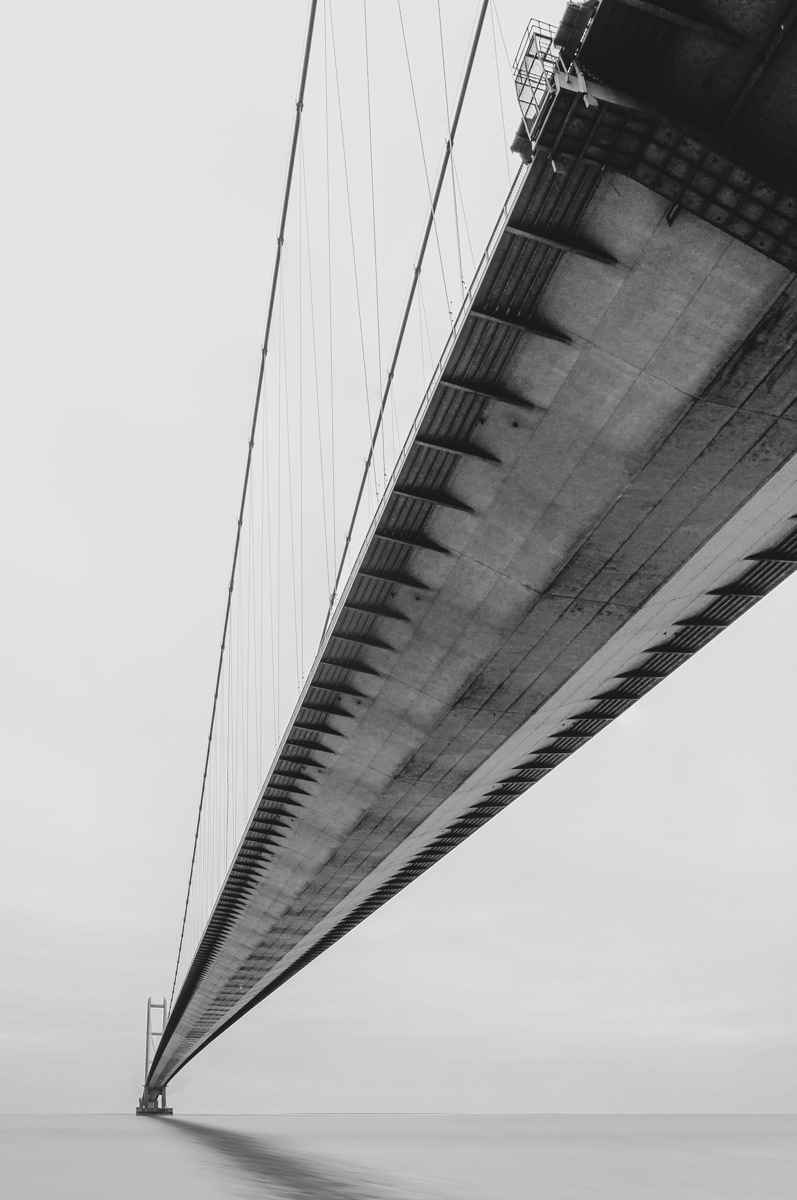 A black-and-white close-up shot of the underside of the Humber Bridge, emphasizing the intricate details of its construction. The bridge stretches into the distance over calm water, creating a striking perspective and showcasing its architectural grandeur against a clear sky.