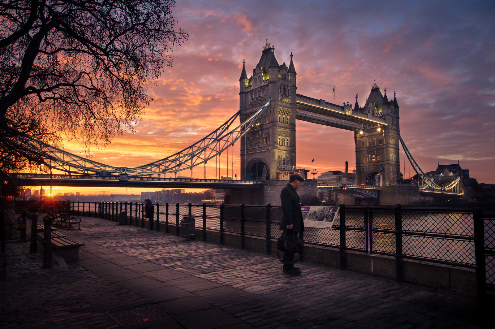 This image captures the Tower Bridge in London at sunrise, presenting a stunning blend of natural and architectural beauty. The sky is painted with vibrant hues of orange, pink, and purple as the sun rises behind the cityscape. The bridge's lights are just beginning to illuminate, creating a mesmerizing contrast with the twilight sky. A lone pedestrian walks along the cobblestone path by the Thames, adding a touch of human element to the serene and picturesque scene. The trees' silhouettes frame the image perfectly, enhancing the overall composition and atmosphere.